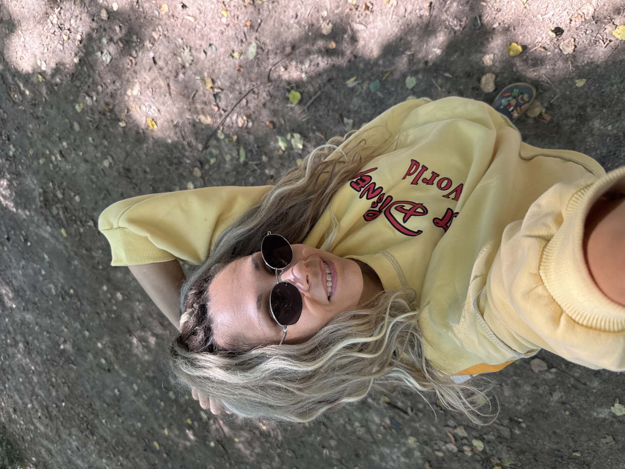 A high-angle, medium shot shows a woman in a yellow Disney World sweatshirt and sunglasses lying on the ground. She is outdoors, with her right arm bent and hand resting on her head, and her left arm holding the phone to take the selfie. The ground beneath her is dirt and scattered leaves, with dappled sunlight filtering through trees overhead. She has blonde, wavy hair and is smiling.