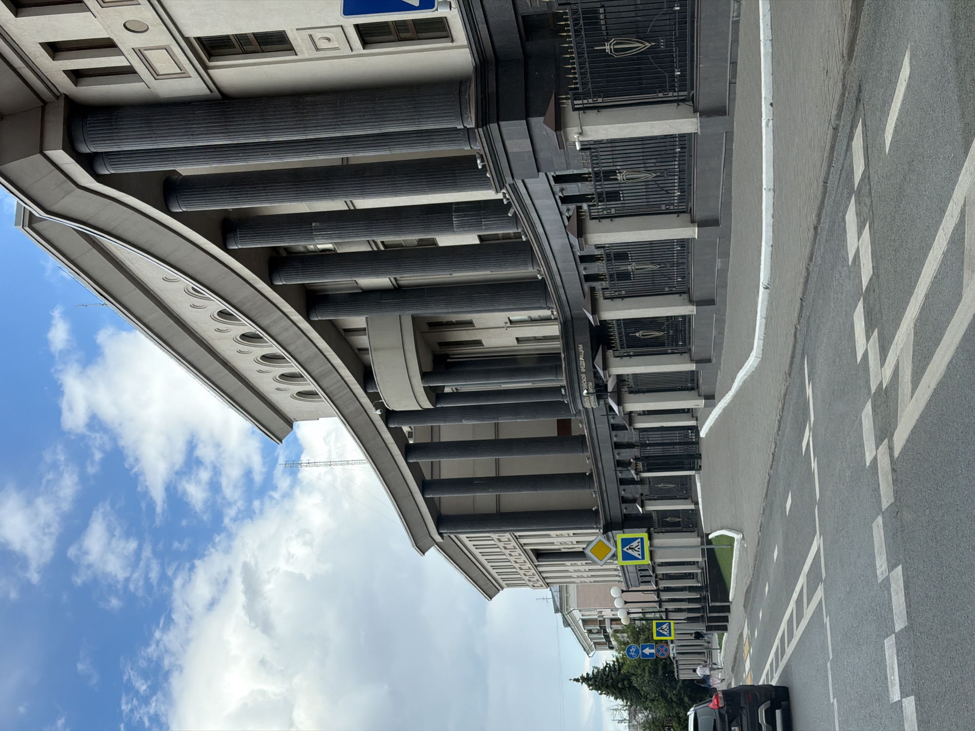 The image depicts a street scene in Kazan, Russia. The camera faces a building with a prominent portico supported by numerous cylindrical columns. Above the portico is a multi-storied facade with windows and decorative elements. A crosswalk is visible, and a car is parked on the side of the road. Various road signs are posted on the side of the building. The sky is cloudy, with patches of blue visible. A tree is visible near the side of the building. The building appears to be in an urban area, potentially near the government buildings of Kazan due to architectural structure and design.