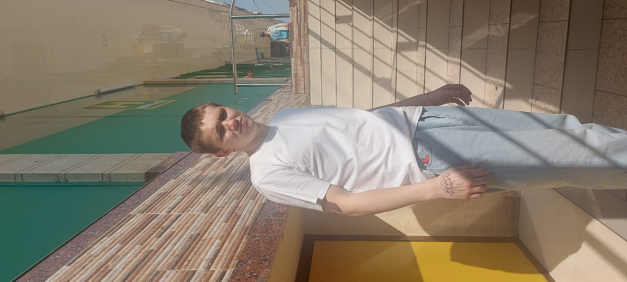 A person lies on a ledge in an urban setting. The ledge is tiled with beige and brown rectangular tiles. The person wears a white t-shirt and light blue denim pants. A hand with a tattoo that resembles a skeleton rests on the ledge. Above the ledge, there is a wall with green and beige horizontal panels. A building is visible in the distance. The location is Tashkent, Uzbekistan. A yellow wall is visible below the ledge. The shadow cast on the background suggests sunlight.