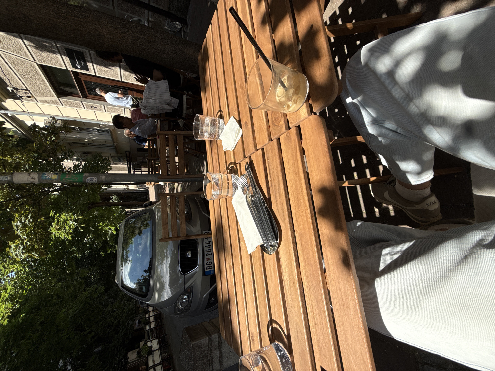 The image depicts a scene at an outdoor cafe in Belgrade, Serbia. A wooden table dominates the foreground, with several empty glasses, napkins, and cutlery placed on it. Part of a person's legs, clad in light blue jeans and Nike sneakers, are visible seated at the table.
Beyond the table, a silver car with the license plate "BG 2444 RI" is parked on the street. In the background, other people are seated at tables, suggesting a busy cafe setting. Buildings and trees line the street, indicating an urban environment. The lighting suggests it is daytime, likely with sunlight filtering through the trees.