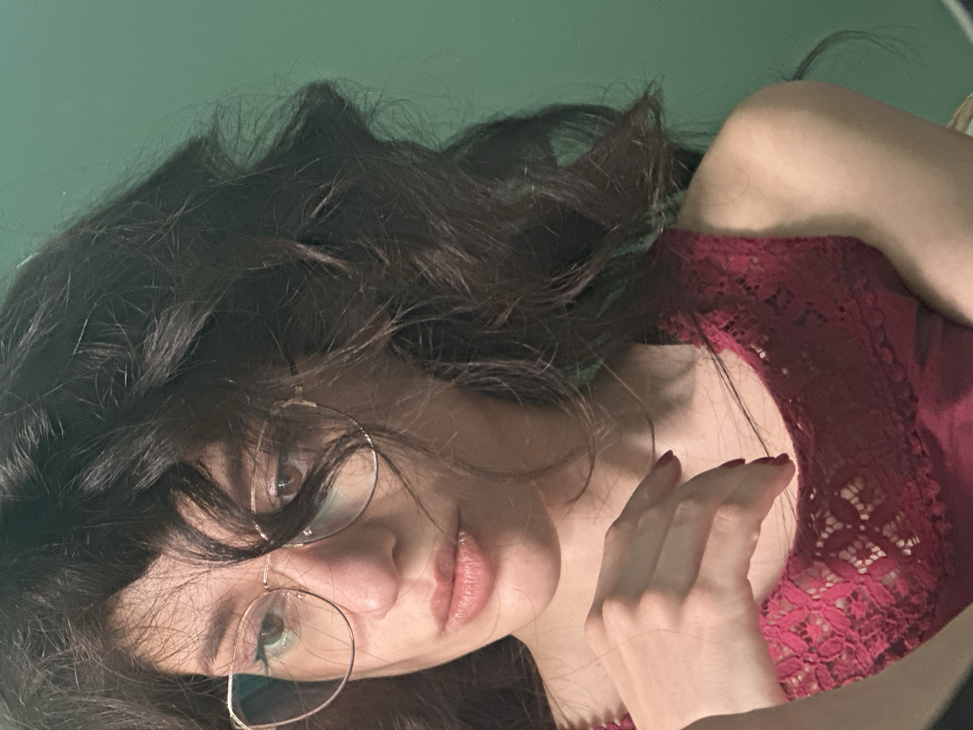 The image is a close-up portrait of a young woman with dark, wavy hair and glasses. She is wearing a dark red lace top. Her left hand is resting on her chest. The background is a solid green wall. The lighting creates shadows on her face, highlighting her features. The image appears to be taken indoors, in a domestic setting. No specific details are present to indicate the location is Sofia, Bulgaria.
