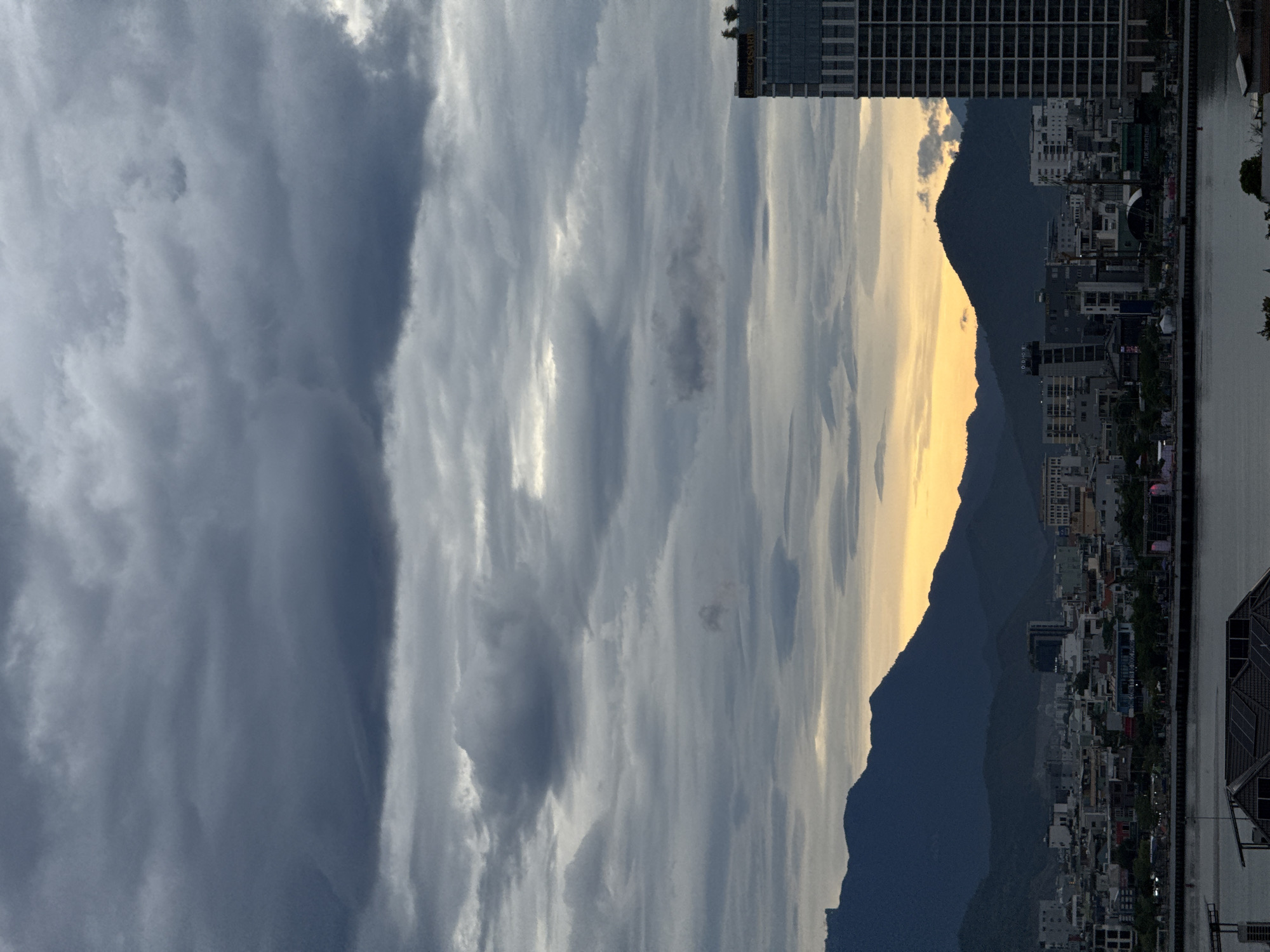 The image captures a city skyline at dusk, with a prominent skyscraper on the right. The sky is filled with dramatic, dark clouds, suggesting an impending storm or the aftermath of one. In the distance, a range of dark, silhouetted mountains forms a backdrop to the city. A narrow band of warm, yellow light is visible between the mountains and the horizon, indicating the setting sun peeking through the clouds. The city itself is a mix of low-rise buildings and taller structures, with lights beginning to twinkle as night approaches. A body of water, possibly a river or bay, lies in the foreground, with some structures visible along its edge. The presence of the One Opera Hotel, a distinctive building with a façade featuring a series of vertical lines and signage at the top, can be identified on the right side of the image, suggesting the location is within the city of Đà Nẵng, Vietnam. There are no people or discernible actions visible in the scene, which is a static view of the urban landscape under a brooding sky.