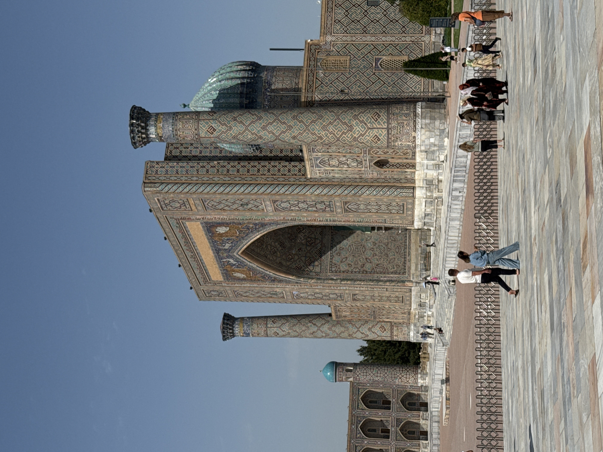 The image depicts the exterior of the Bibi Khanym Mosque in Samarkand City, Uzbekistan. The architecture features tiled mosaics, high arches, tall minarets, and a large dome. People are walking across the paved courtyard of the mosque, and a metal fence separates the building from the walkway. The sky is blue.