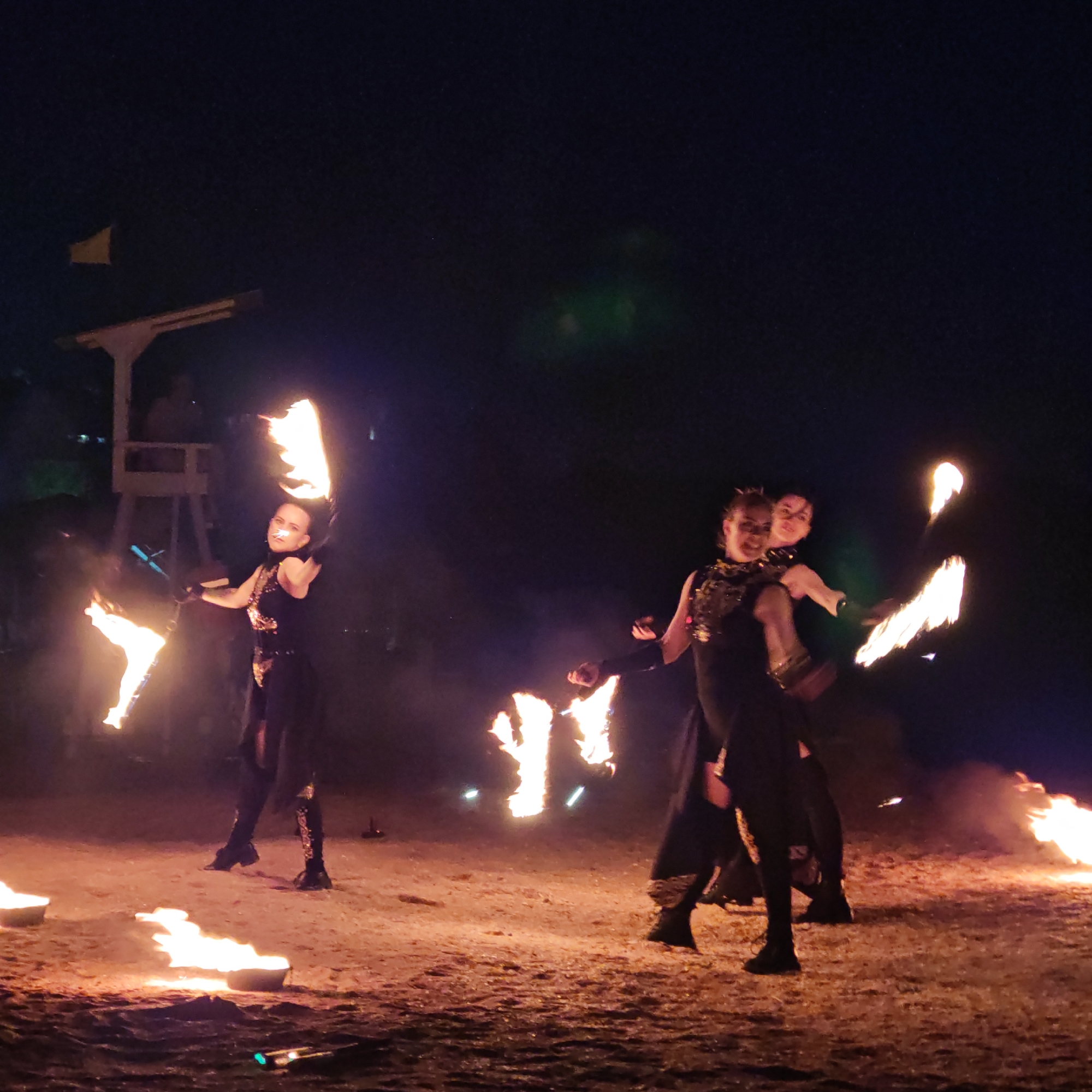 In the city of Shcholkine, Ukraine, a group of performers engage in a fire-dancing spectacle at night. Three individuals, two women and a third person whose gender is obscured, are the central figures. The woman on the left, wearing a black costume adorned with gold sequins on the bodice and patterned leggings, holds flaming props in both hands, with a large flame emanating from her right side. To her right, two individuals are positioned closely together. The woman in front, also in a black, sequined costume, is seen from the side and slightly behind, with her arms outstretched, holding a flaming prop. Behind her, another person's face is visible, looking towards the viewer, with a flaming prop also in their hand.
The performance takes place on a sandy or earthy surface, illuminated by the bright flames and the glow from portable fire bowls scattered on the ground. In the background, a wooden structure resembling a lifeguard tower or observation post stands, with a yellow flag visible on top. The dark night sky forms the backdrop, with hints of ambient light and some blurred green and purple hues suggesting artificial lighting or atmospheric effects. The scene captures a moment of dynamic movement and intense visual energy from the fire performance.
