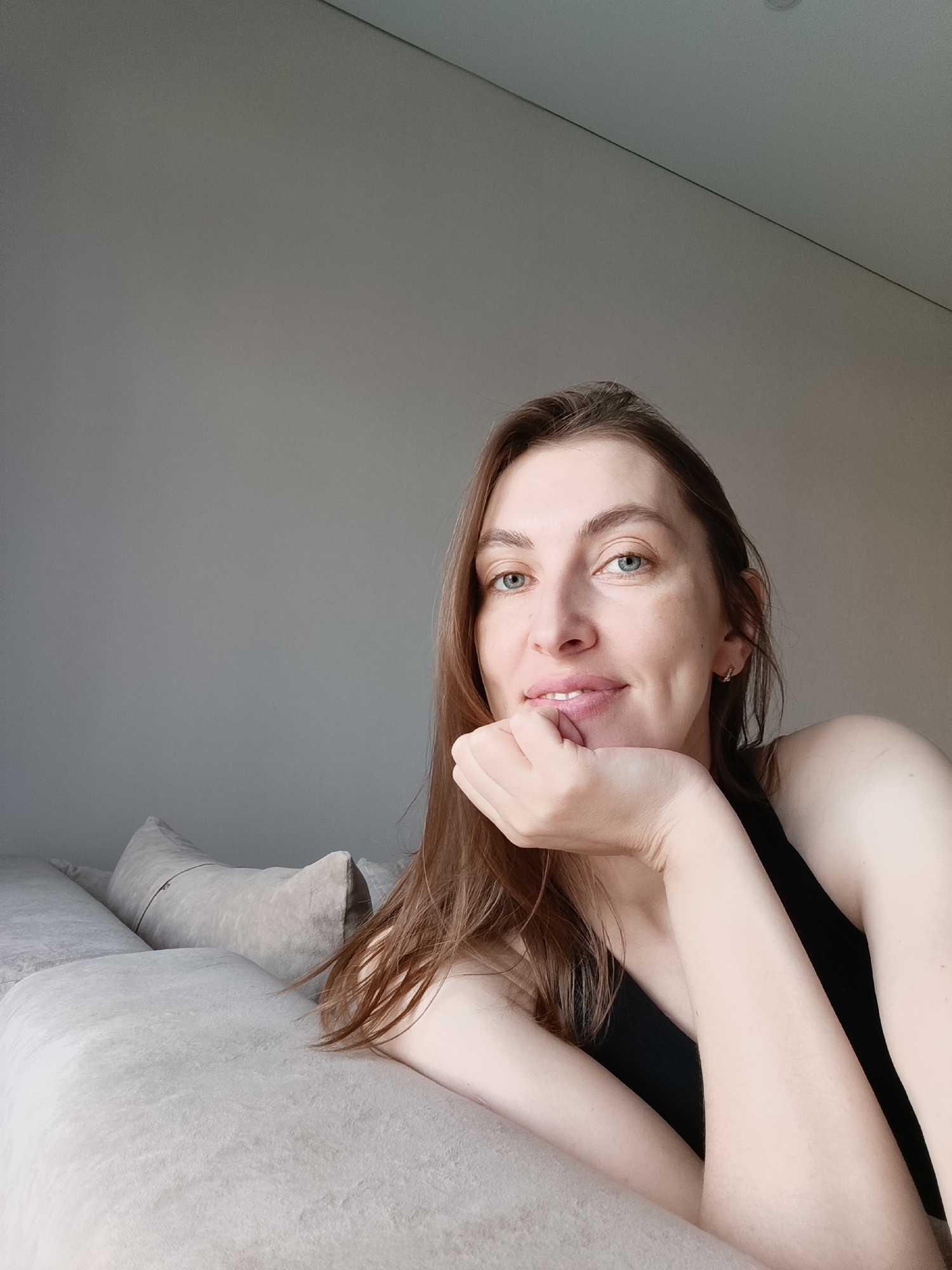 A woman with light brown hair and blue eyes is lying on a light-colored couch with several pillows behind her. She is wearing a black tank top and her left hand is supporting her chin. The wall behind the couch is light gray and appears to be part of a room with an angled ceiling. The location is Chaikovsky, Russia.