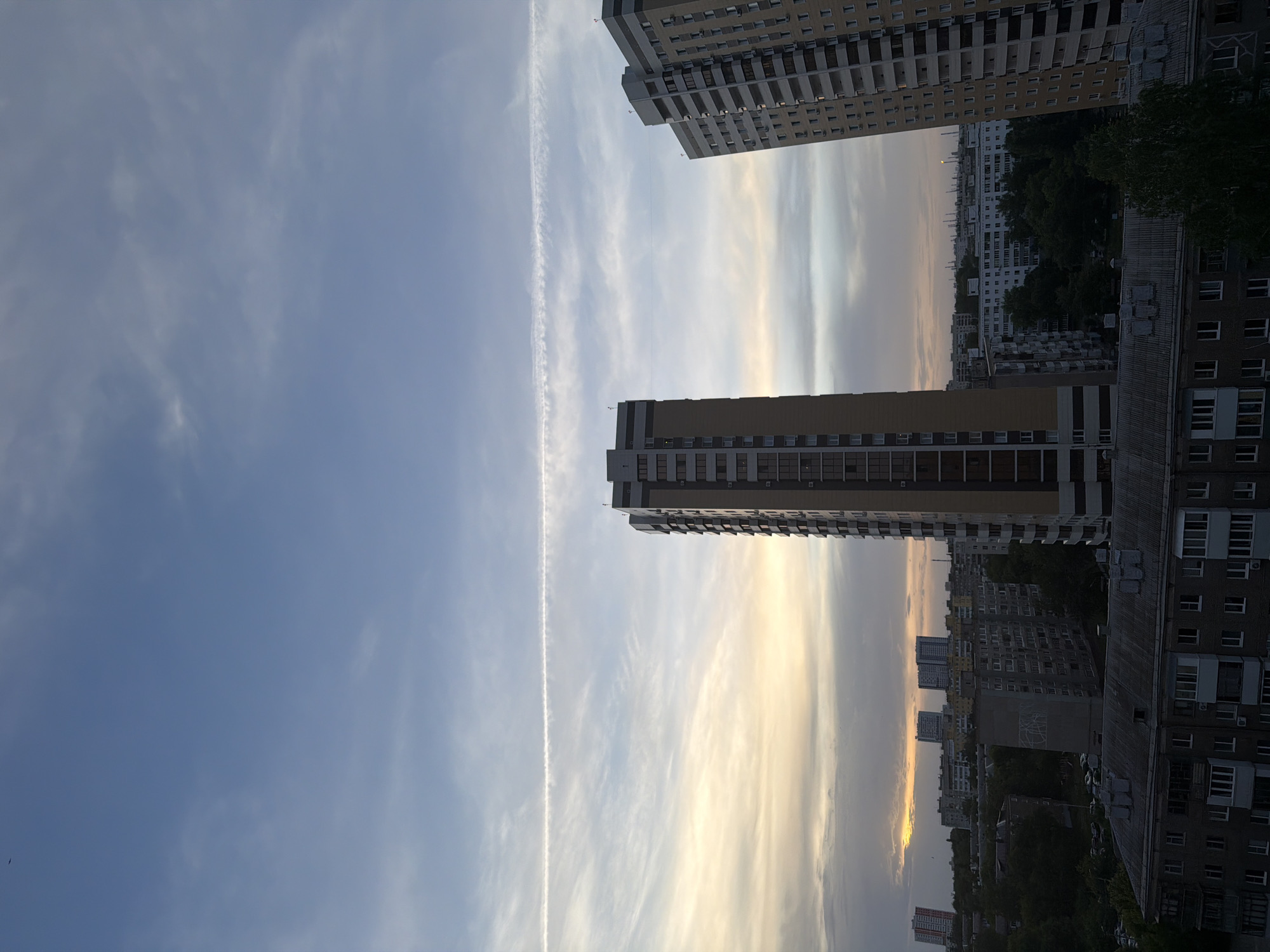 The image shows a cityscape with several tall buildings. The sky is visible, with streaks of clouds and a contrail from a jet. The buildings are a mix of older, shorter structures and newer, taller apartment blocks. The scene is likely taken during either sunset or sunrise based on the coloring of the sky.