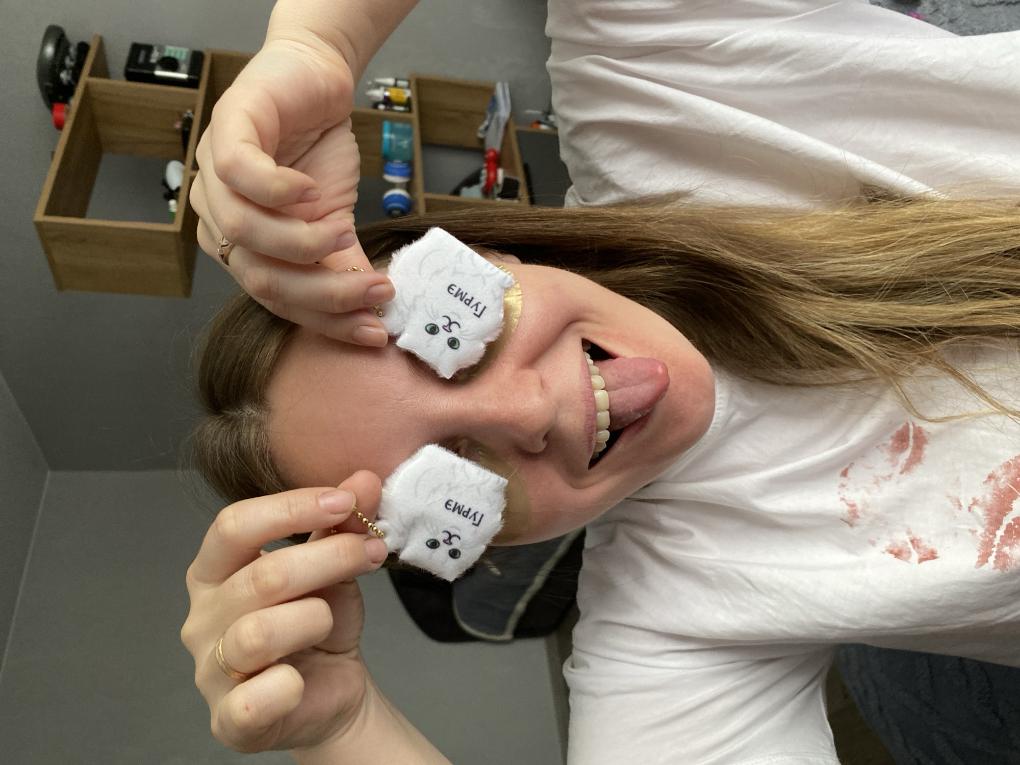A woman in a white t-shirt is upside-down. She is holding two white, square-shaped cat plush toys with Cyrillic writing near her eyes. She is sticking out her tongue. There is a shelf in the background. The t-shirt has a red stain on it. The image may be from Noviy, Russia, but there is no way to confirm it from the visual data provided.