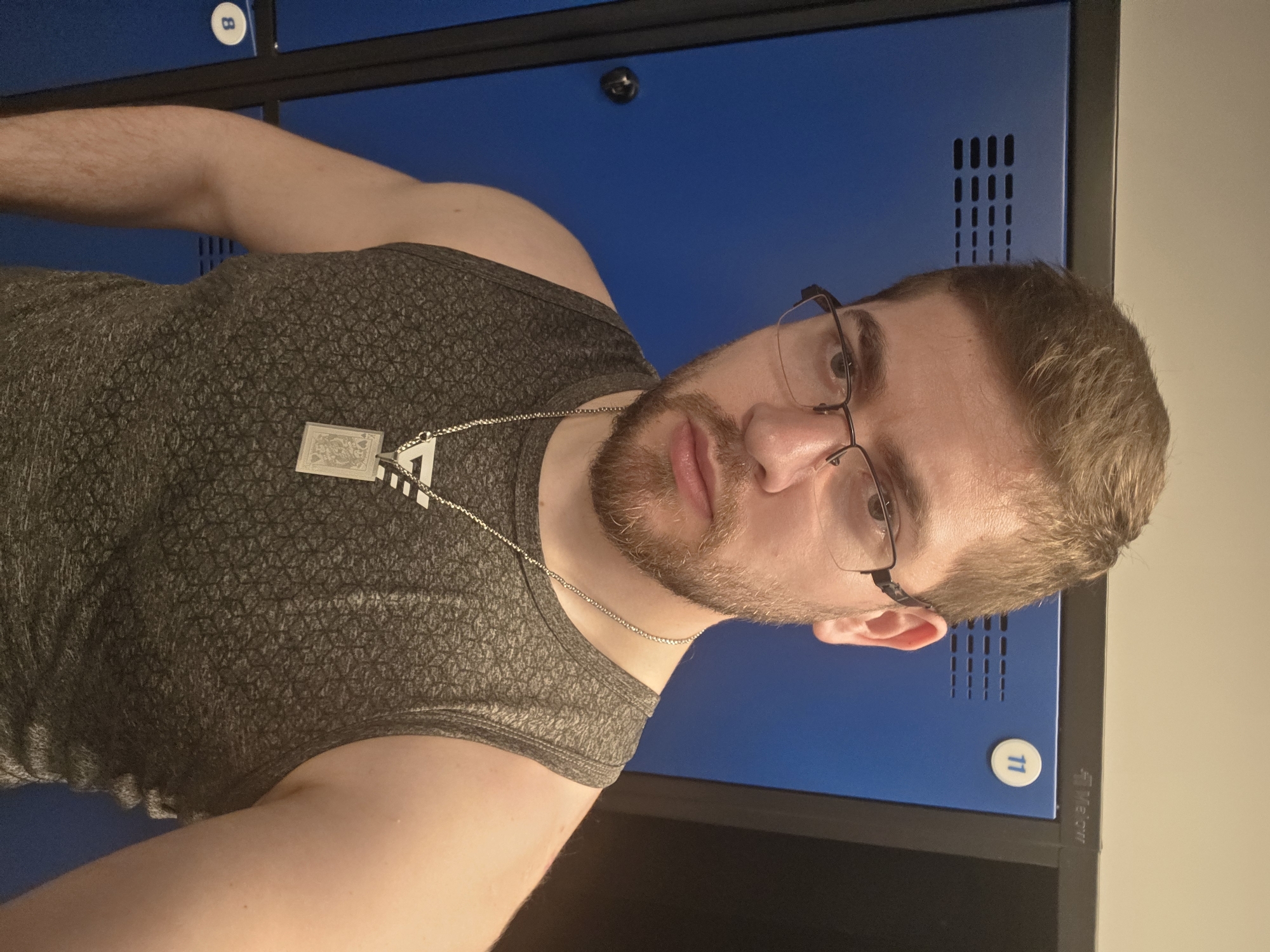 A fair-skinned man wearing glasses, a gray patterned tank top, and a silver necklace is looking at the camera. He has short brown hair and a reddish beard. Behind him are blue lockers with ventilation slots and number tags, possibly in a gym or changing room. The specific location within the city of Plovdiv, Bulgaria, is not identifiable from the image.