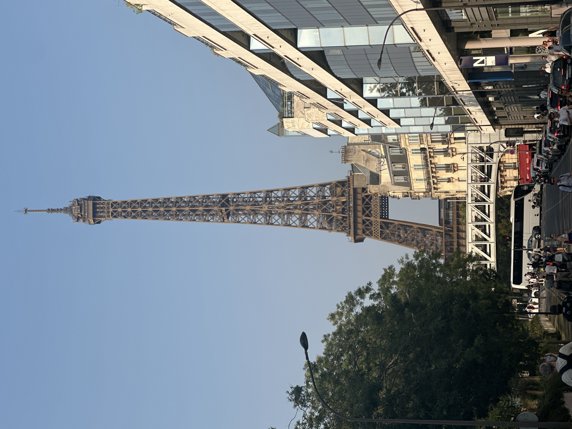 The image depicts a view of the Eiffel Tower in Paris, France, from a street level perspective. In the foreground, there is a sidewalk with people walking, a street with cars, and a white bus. On the right side of the image, there is a modern building with a glass facade. On the left side, there are trees and a lamppost. The Eiffel Tower is visible in the background, towering over the surrounding buildings. The sky is clear and blue.