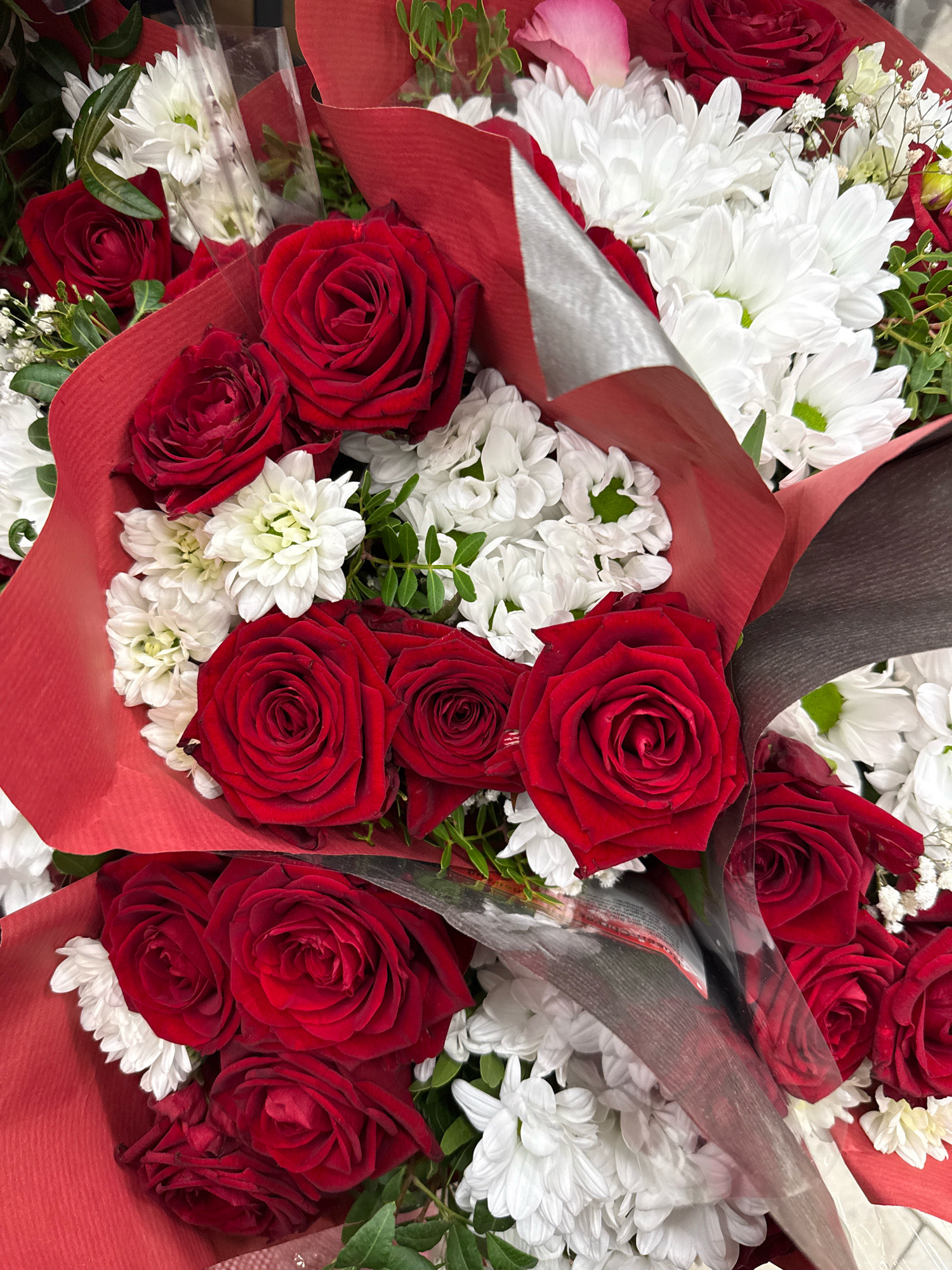 The image shows a close-up view of several bouquets of flowers, predominantly red roses and white chrysanthemums. The flowers are artfully arranged and wrapped in red and silver paper. The lighting suggests an indoor setting, possibly a flower shop or a market stall. There are no people or animals visible in the image, and no discernible details of the city of Göttingen, Germany, are present. The focus is entirely on the floral arrangement.