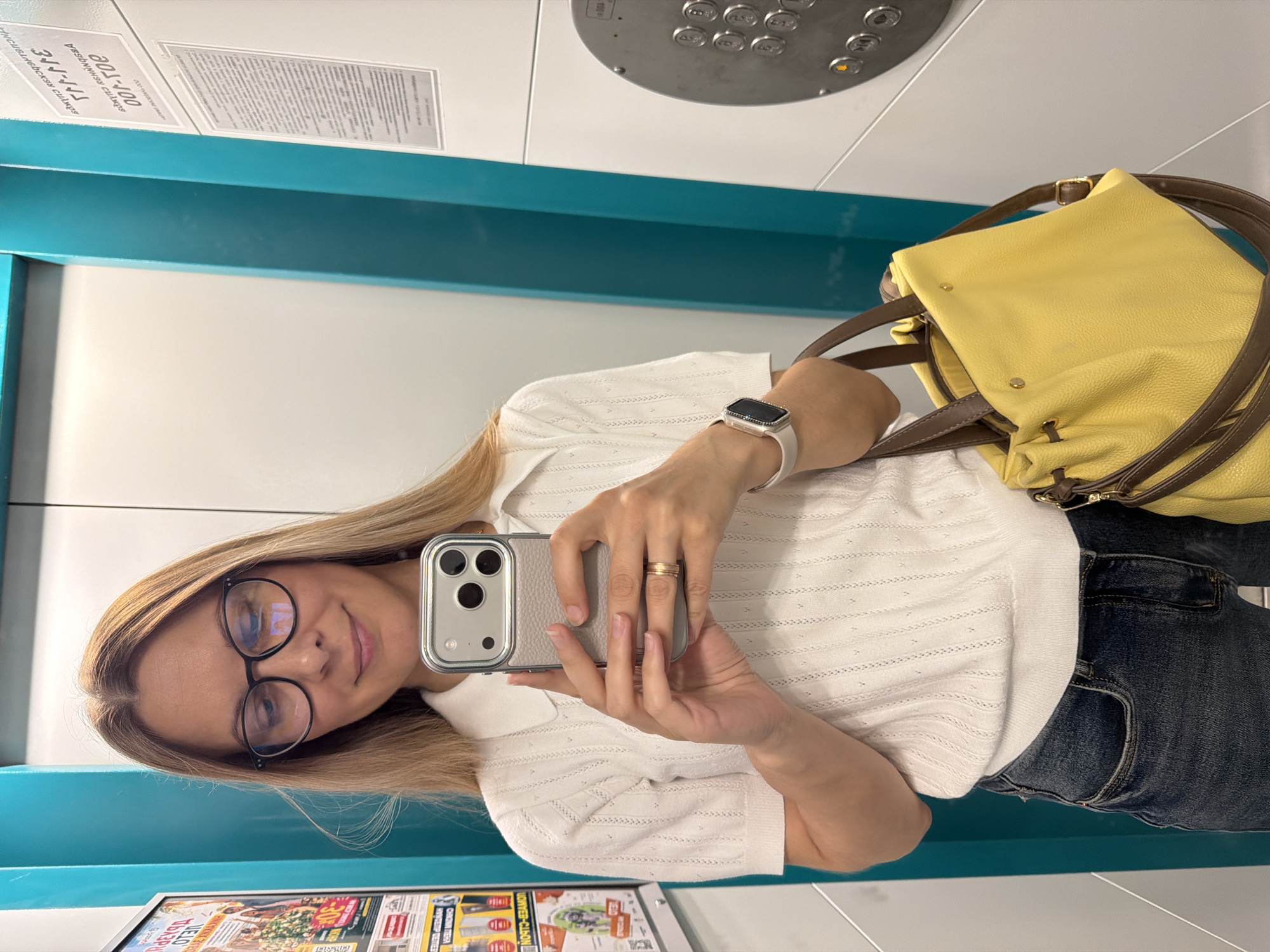 In Izhevsk, Russia, a woman stands in an elevator, taking a selfie. She is wearing a white short-sleeved polo shirt with a textured knit pattern and dark blue jeans. She has long blonde hair, wears black-rimmed glasses, and has on a smartwatch with a white band and a ring on her right hand. She is holding a smartphone in her right hand, pointed towards a mirror. A bright yellow handbag with brown leather straps rests on her left hip. The elevator walls are light grey and teal, with a metallic panel on the right side displaying elevator buttons. Posters are visible on the left wall of the elevator.
