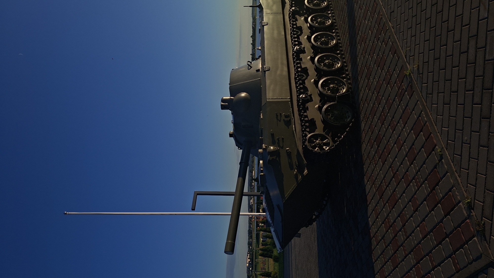 A weathered, dark green tracked vehicle, resembling a military tank, is positioned on a paved area. The paving consists of interlocking rectangular blocks in shades of red and gray, laid in a pattern. The vehicle's tracks are clearly visible and appear to be in good condition. Its main gun barrel is extended forward. In the background, a tall, thin metal pole stands vertically, and a thin dark gray vertical structure extends from the ground. Beyond this, a body of water is visible, with a distant treeline and buildings on the opposite shore. The sky above is a clear, deep blue, with a faint crescent moon visible. The lighting suggests it is late afternoon or early evening, with long shadows cast by the vehicle and the pole. The location is identified as Smalyavichy, Belarus. There are no people or animals visible in the image.