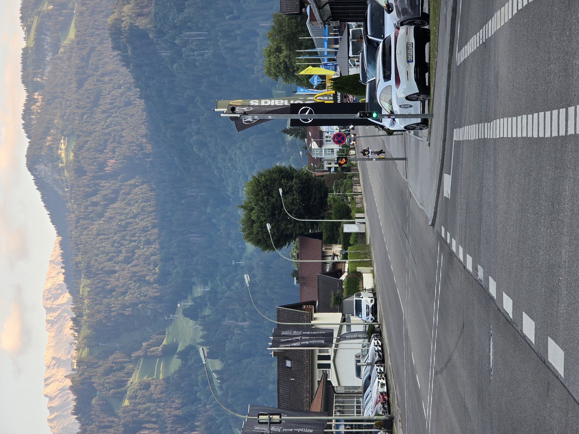 The image depicts a roadside scene in Garmisch-Partenkirchen, Germany, with a backdrop of lush, tree-covered mountains and a rugged cliff face bathed in the warm light of either sunrise or sunset. The foreground is dominated by a paved road with white lane markings and a pedestrian crossing. Parked cars are visible along the roadside, including several Mercedes-Benz vehicles, as indicated by the text "Mercedes-Benz Junge Sterne" on banners. A white Mercedes-Benz with license plate "MIR 6373E" is prominently displayed. Traffic lights show a green signal, and a "no entry" sign is also present. In the distance, ski lifts ascend the mountains, suggesting this is a location popular for both summer and winter recreation. The overall atmosphere is serene and picturesque, capturing the beauty of the Bavarian Alps.