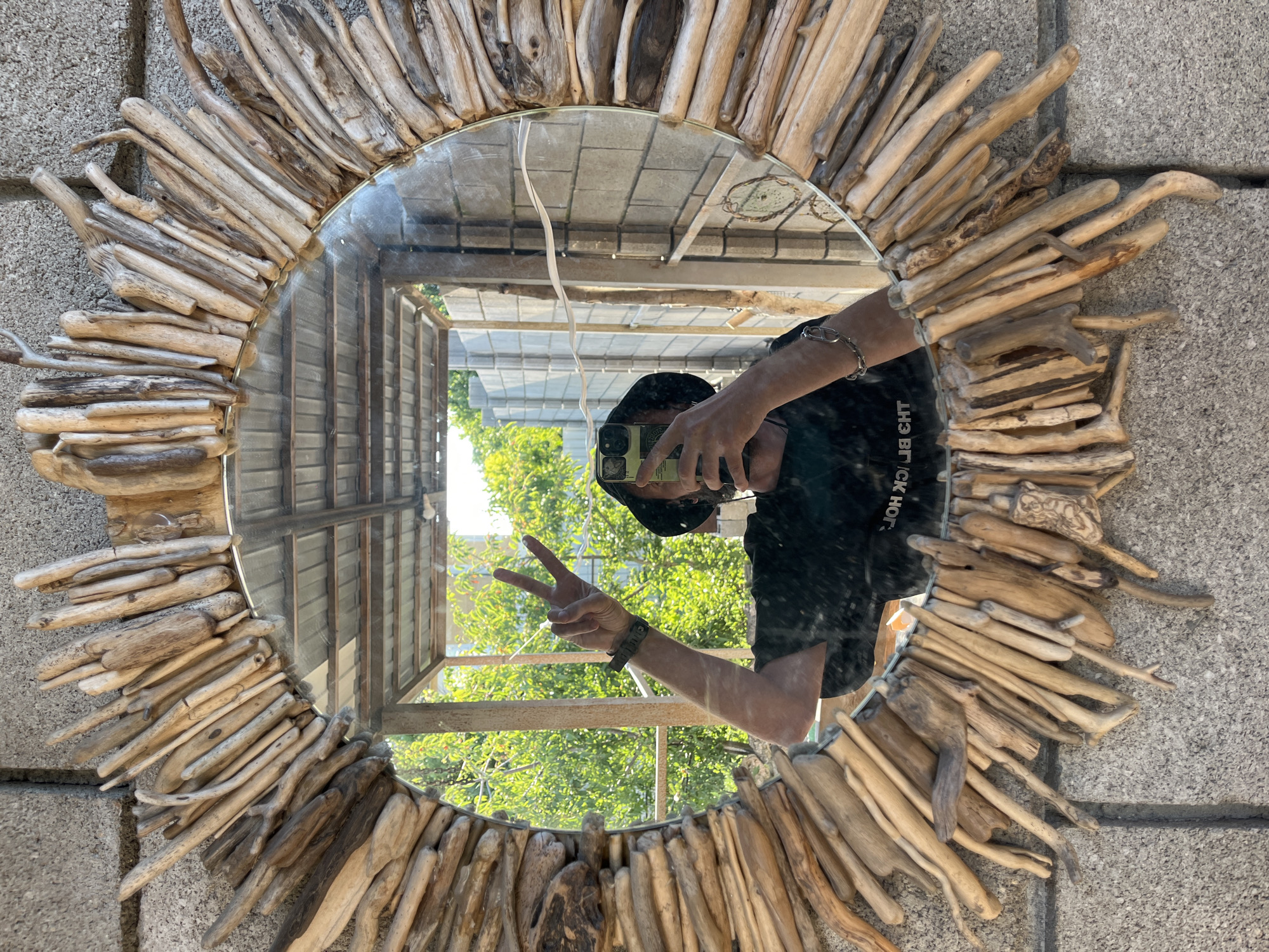 A person wearing a dark hat and dark t-shirt is reflected in a round mirror. The mirror is framed with a sunburst-like arrangement of natural driftwood pieces of varying sizes and shades of brown and grey. The person is holding a smartphone in their right hand, angled to capture the reflection. Their left arm is raised, and they are making a peace sign with two fingers. The background visible in the mirror shows a concrete block wall and lush green trees. The mirror is mounted on a textured grey concrete surface.