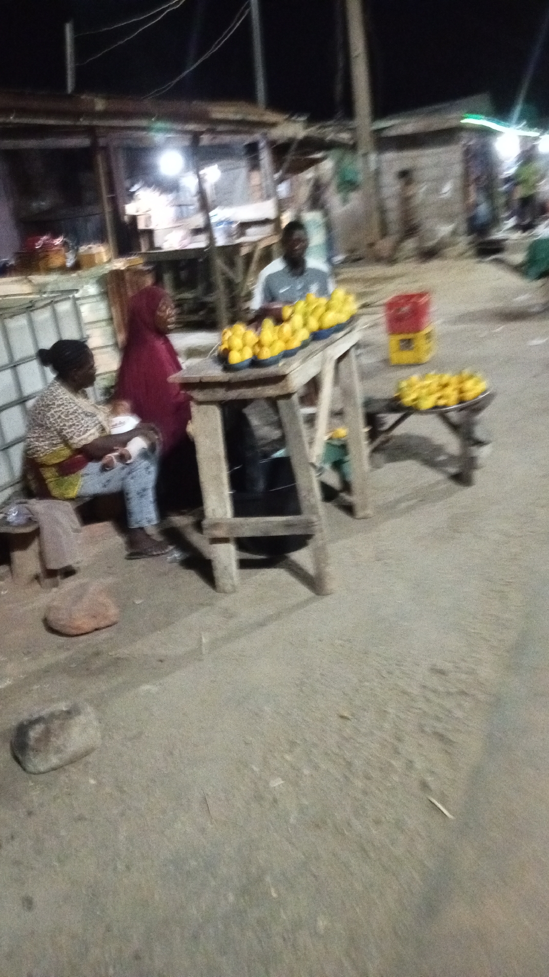 The image shows a street scene at night in Zango-ndaji, Nigeria. A man stands behind a wooden table covered with yellow fruits, likely selling them. Two women are seated on a bench near the table. One of the women, dressed in a red hijab, appears to be breastfeeding a baby. A red and yellow crate sits nearby, along with a smaller table also displaying fruit. The ground is paved. Buildings are seen in the background, illuminated by artificial lighting.