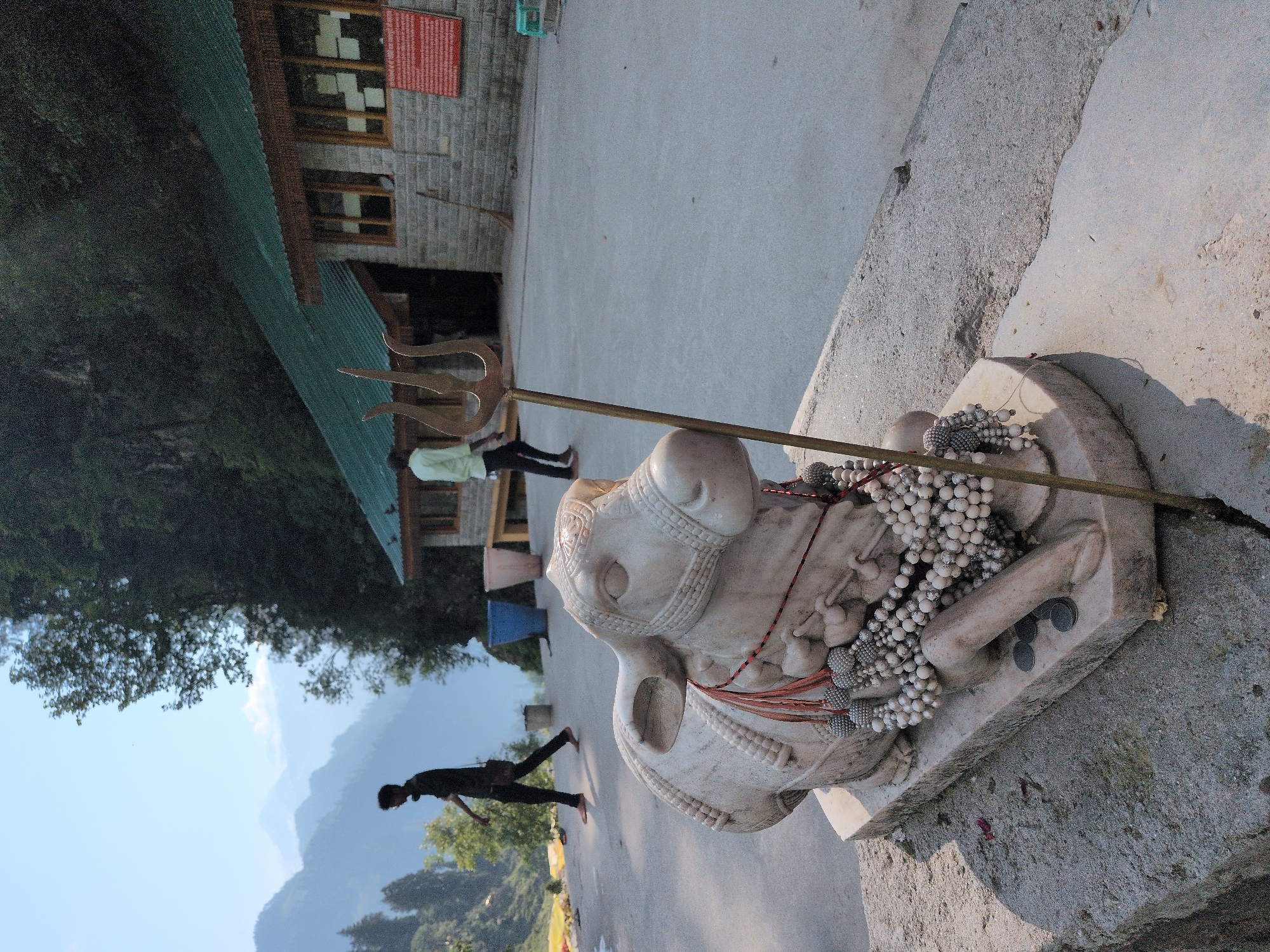 This image depicts an outdoor scene near a building with a green roof, possibly in the city of Majhach, India. In the foreground, a white marble statue of a cow is prominently displayed, adorned with strings of white beads and red tassels. A bronze trident is positioned behind the cow statue. Two individuals are visible in the middle ground: one, silhouetted in black, walks away from the camera on a paved surface, while another, wearing a light green shirt, walks towards the building. The background features lush green trees and distant mountains under a clear blue sky. A stone building with wooden window frames and a red sign is visible behind the people and the cow statue. The overall environment appears to be a religious or spiritual site given the presence of the cow statue and trident.