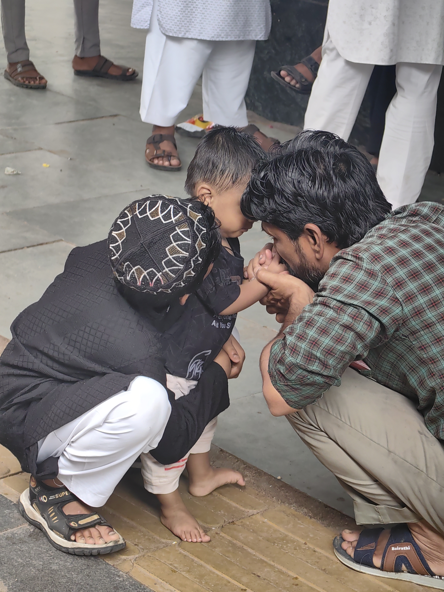 In Khandwa, India, a man in a plaid shirt is tenderly holding the hand of a young child, his face close to the child's. The child, wearing a black cap with a decorative pattern and a black garment, is embracing another child who is also in black attire. Both children have their faces obscured. The scene takes place on a paved outdoor area with a textured yellow and gray patterned ground. Several pairs of feet are visible in the background, some wearing sandals and others bare. The lighting suggests it is daytime.