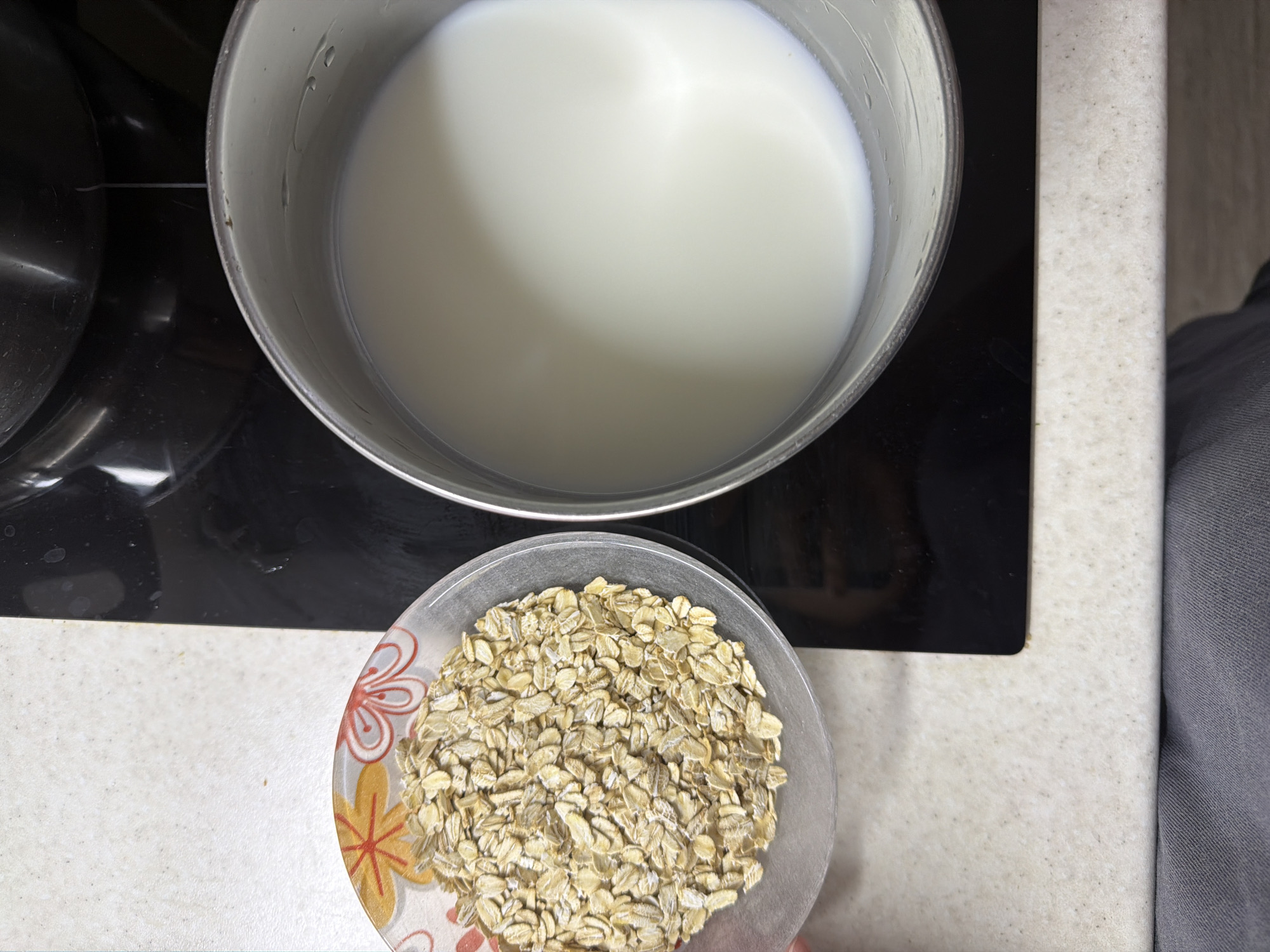 A top-down shot shows a cooking scene, potentially for oatmeal. A stainless steel saucepan is filled with milk and sits on a black stovetop. Next to the saucepan is a clear glass bowl filled with dry rolled oats. The bowl has a floral design. A person wearing gray clothing is standing beside the counter. The location is Chaikovsky, Russia.