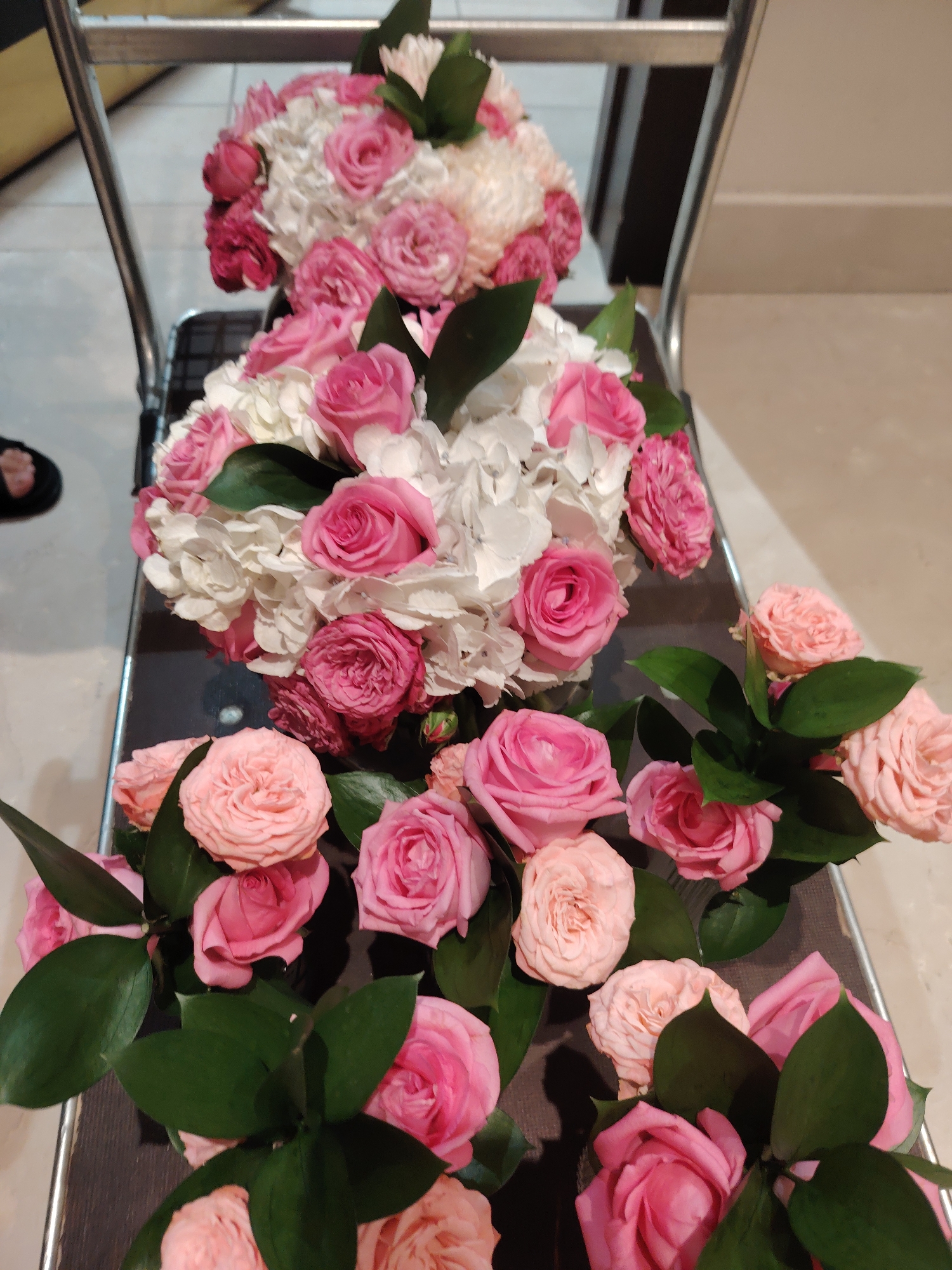 The image shows a collection of floral arrangements, predominantly pink and white roses, with some interspersed white hydrangeas and dark green foliage. The flowers are arranged in bouquets and are positioned on what appears to be a dark-colored, tiered stand or chair. The background is blurred but suggests an indoor setting with muted lighting and possibly a tiled floor. No specific landmarks of Konigil, Uzbekistan, are visible. There are no people actively interacting with the flowers in the frame.