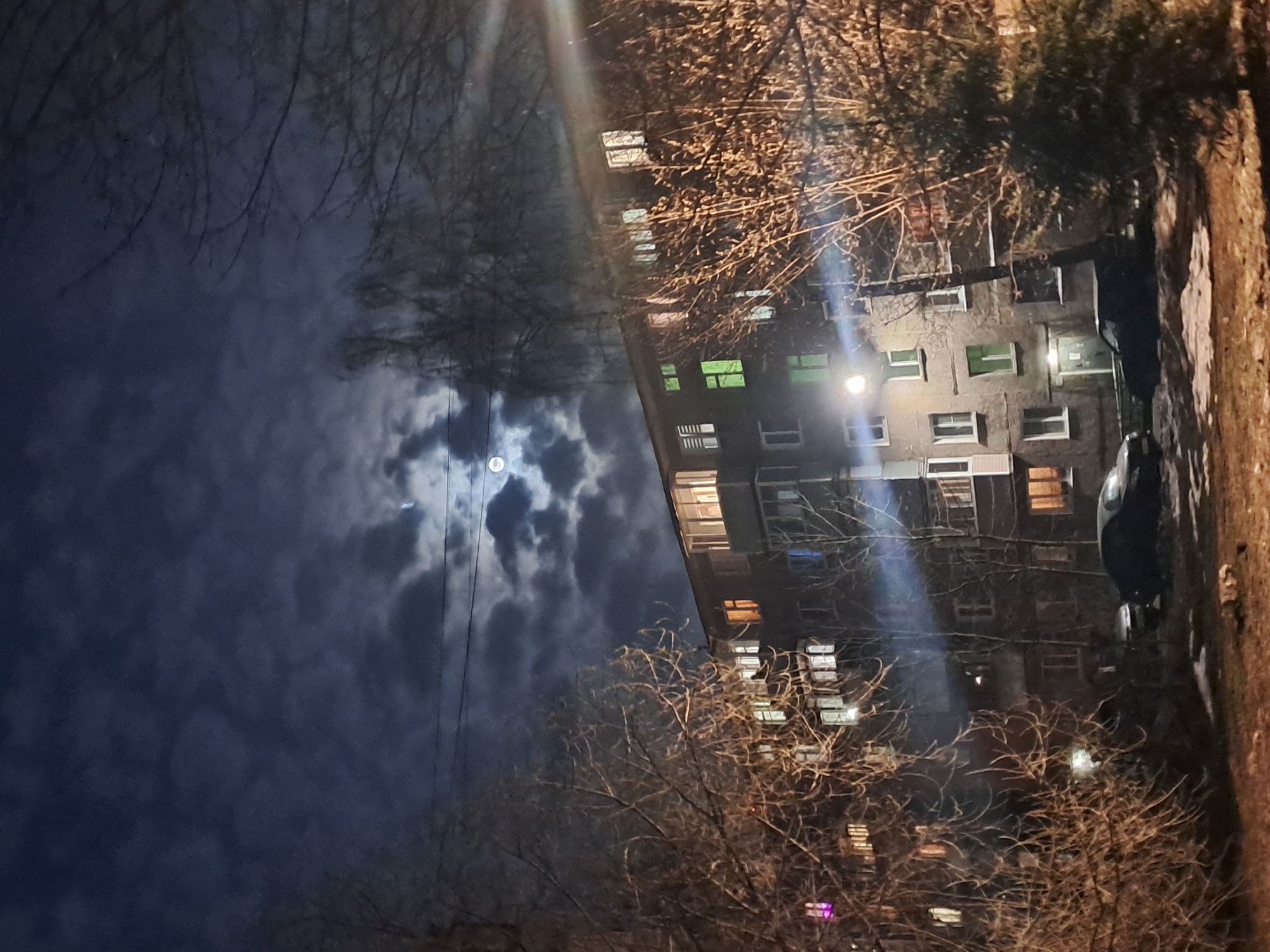 The image, taken at night in what appears to be Elektrostal, Russia, depicts a multi-story residential building partially obscured by bare trees. The building is illuminated by both interior lights in some windows and a bright street lamp. A vehicle is parked next to the building. In the upper-left quadrant, the moon is visible through a layer of clouds. A power line also runs vertically through the sky. A sidewalk/green patch is visible on the right edge.