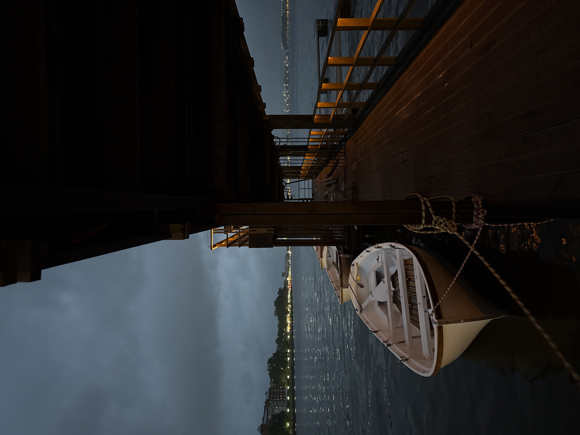 This nocturnal image, captured in Votkinsk, Russia, displays a covered wooden dock extending over a body of water under a dark, overcast sky. In the foreground and midground, three white rowboats with beige undersides and wooden interiors are moored to the dock's side, with a prominent rope securing the closest boat to a wooden post. The dock features a wooden plank floor and wooden railings, illuminated by warm, yellowish artificial strip lights integrated into the top rail, casting reflections on the damp surface of the deck.
To the left, the dark water exhibits ripples and reflections from distant city lights. The opposing shoreline, visible across the water, presents a cityscape with numerous illuminated buildings and trees, indicating a populated area. The overall scene is devoid of human presence. The primary light sources are the dock's artificial lighting, the distant city lights, and the minimal ambient light from the dark sky, suggesting twilight or night conditions.