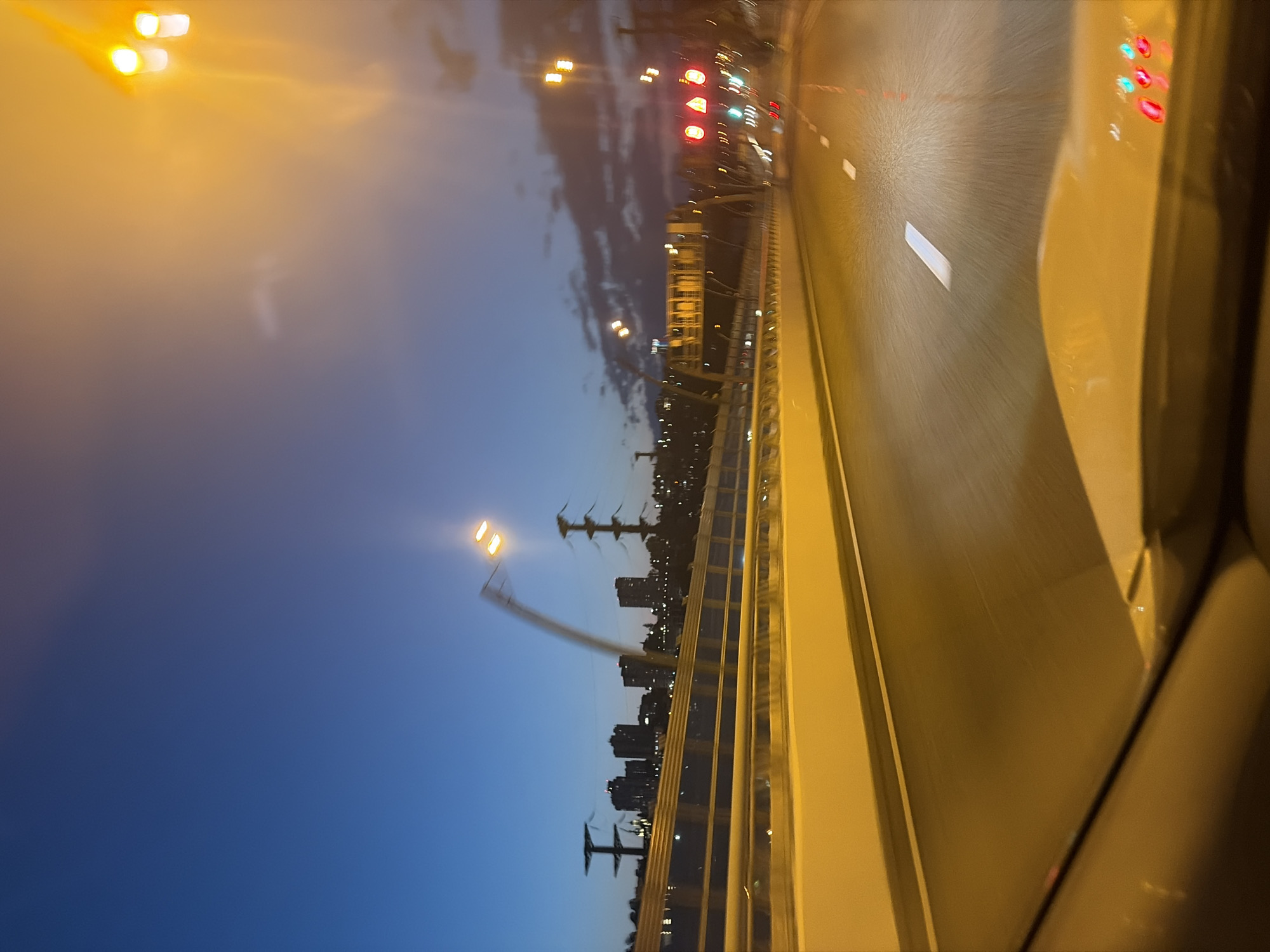 The image depicts a view from the driver's seat of a vehicle traveling on a highway at dusk. The sky is a deep blue transitioning to orange at the horizon, with scattered clouds. On the left side of the highway, a yellow barrier separates the road from the embankment, which is lined with trees and silhouetted buildings of a city. Several tall structures, possibly power line towers, are visible. The highway itself has white lane markings, and the road surface appears wet. On the right side of the road, there are streetlights and traffic lights, some of which are illuminated. The foreground shows the dashboard and windshield of the car, with reflections of the lights on the glass. The image is taken with a motion blur, suggesting the vehicle is in motion. This scene is consistent with a view of the highway leading into or out of Sochi, Russia, particularly with the juxtaposition of the highway infrastructure, urban skyline, and the natural elements of the sky and vegetation common in the region.