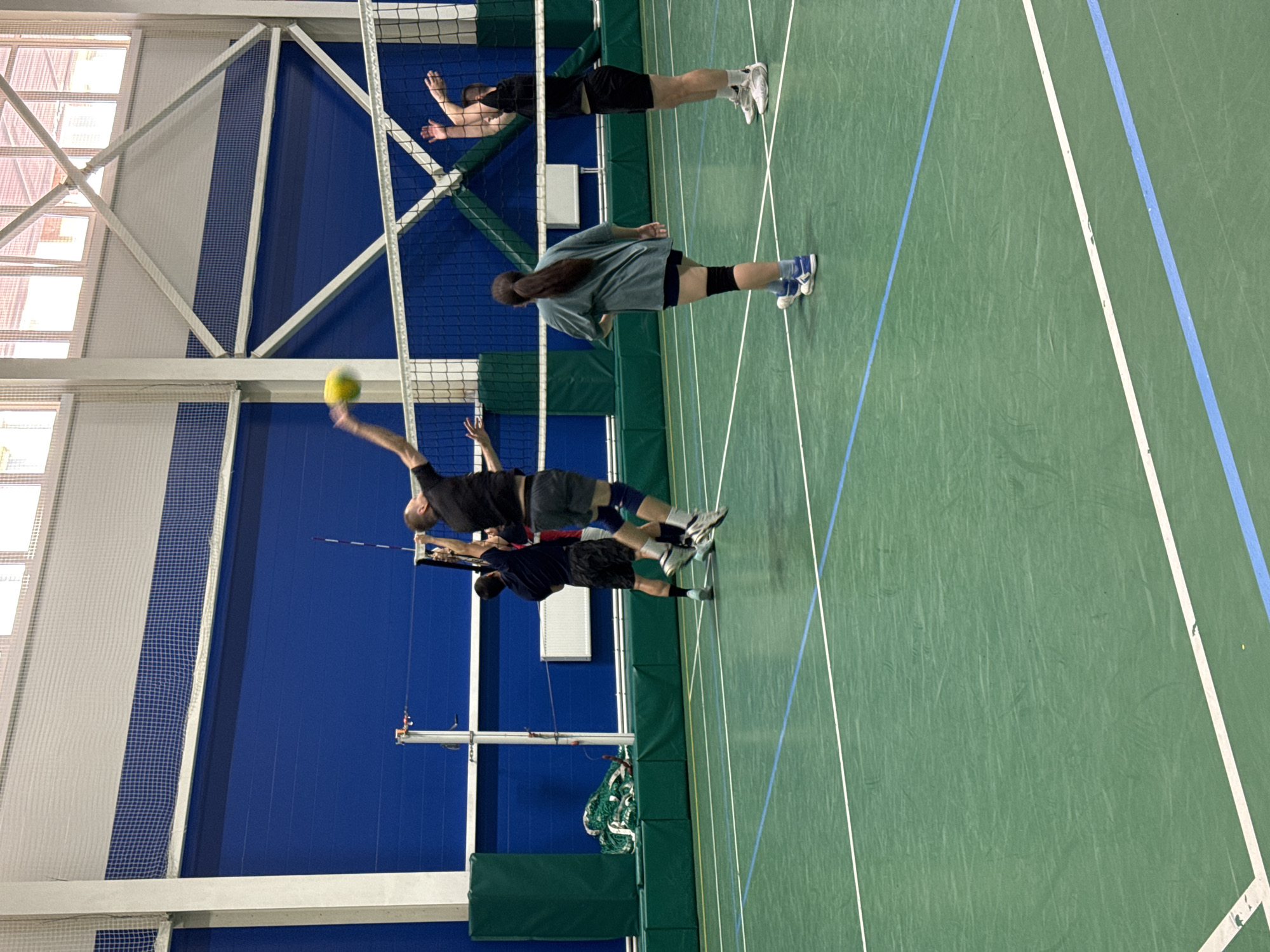 The image depicts an indoor volleyball game taking place on a green court with white and blue boundary lines. The court is within a gymnasium with a high ceiling and blue walls. There are five individuals visible in the scene. Two men are in mid-air, jumping to play the ball. One man, wearing black shorts and a black t-shirt, is at the peak of his jump, reaching to hit a yellow volleyball. Another man, in dark shorts and a dark t-shirt, is also jumping near the net. Two other individuals, one man and one woman, are on the court, observing the play. The man is wearing dark shorts and a black t-shirt, while the woman is wearing dark shorts, a grey t-shirt, and knee pads. The net is stretched across the middle of the court. In the background, there are green padding along the wall, a volleyball net rolled up, and white radiators. The lighting suggests an indoor setting, possibly a sports complex. The scene is consistent with a recreational or amateur volleyball match. Based on the provided information, this image was taken in the city of Saint Petersburg, Russia.