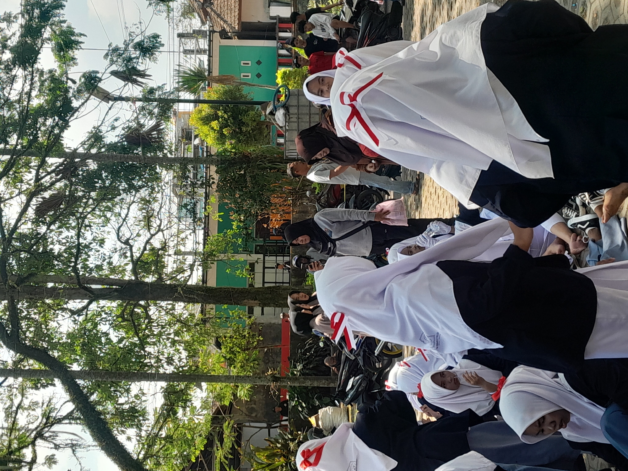 The image depicts a gathering of people outdoors in what appears to be a street or courtyard area. Several individuals, predominantly young women wearing white hijabs with red ribbon accents, are the central focus. Some are seated on the ground, while others are standing. In the background, there are motorcycles parked, and buildings with green and white facades are visible, suggesting an urban or semi-urban setting. Trees with lush foliage are present, providing shade and contributing to the outdoor atmosphere. The presence of flags or banners, indicated by the red and white ribbons on the hijabs, hints at a possible event or celebration. The overall scene suggests a casual social gathering or a specific organized event in Genteng Kulon, Indonesia.