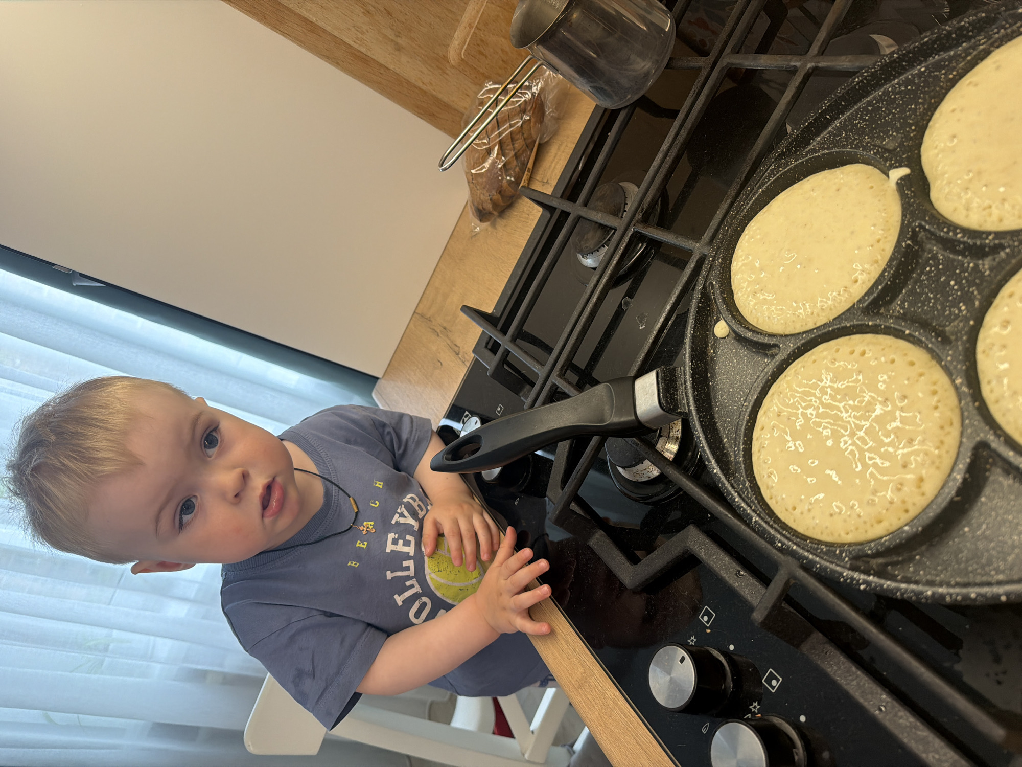 A young child stands next to a stovetop with a pancake pan containing several cooking pancakes. The child is wearing a gray t-shirt with a sports design, a necklace, and holding a yellow and green ball. A pot and loaf of bread sit on the stovetop behind the pancake pan. The countertop and surrounding walls are light colored. It is not possible to confirm the location is Votkinsk, Russia from the image.