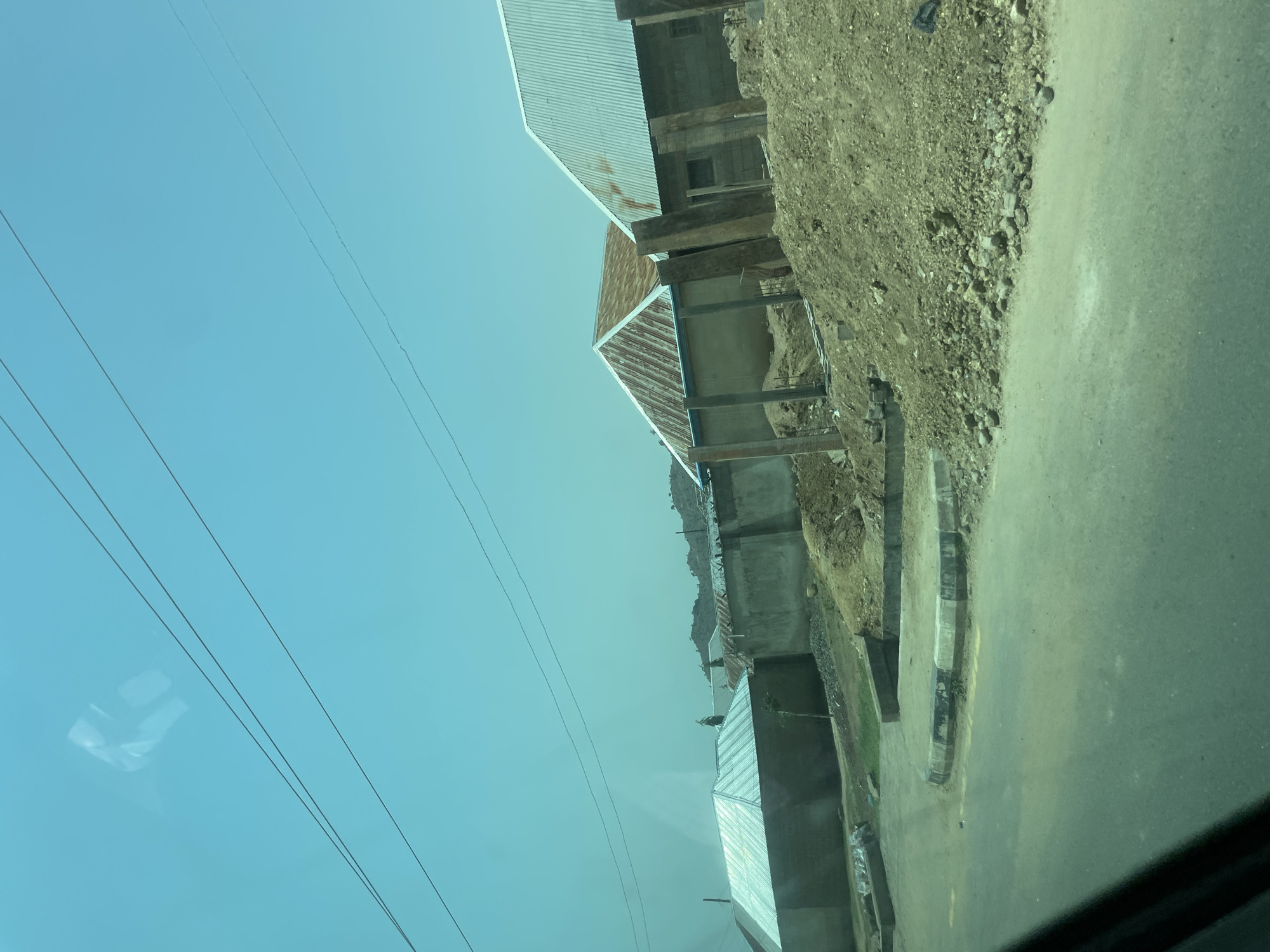 This image captures a street scene in Bauchi, Nigeria, characterized by a clear, bright blue sky and several residential buildings. The foreground shows a paved road with a raised curb, suggesting a developed area within the city. In the mid-ground, a row of houses with corrugated metal roofs are visible, some appearing partially constructed or under renovation, evidenced by piles of dirt and scaffolding-like wooden posts. A concrete wall separates these buildings from the road. The background reveals more buildings and a slightly hilly landscape, indicating the undulating topography of Bauchi. The overall impression is of a typical urban or semi-urban environment in Nigeria, with ongoing construction and residential activity.