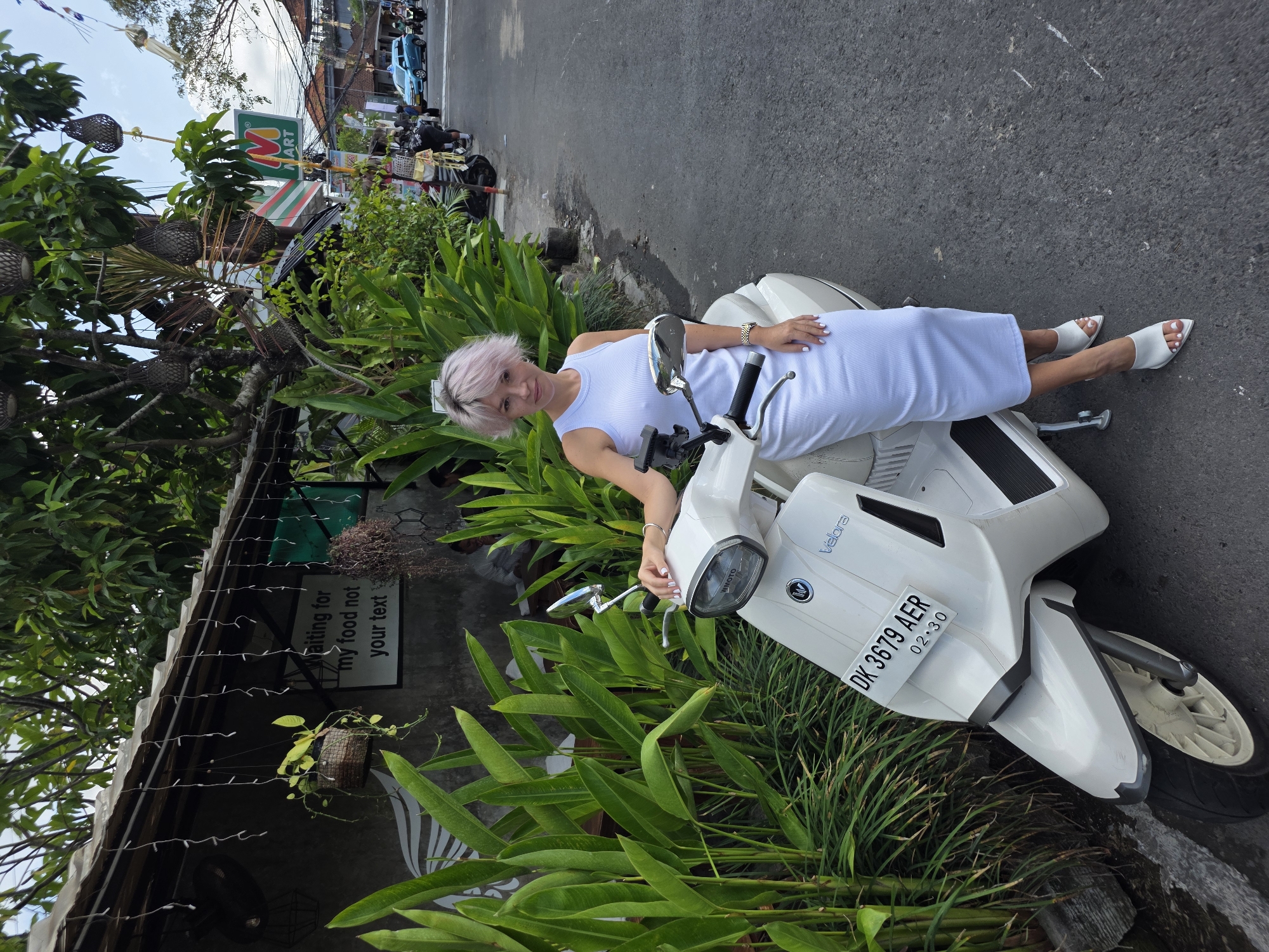 A woman with short pink hair stands next to a white scooter on a street in North Kuta, Indonesia. She is wearing a white sleeveless dress and white pointed-toe mules. The scooter has a license plate that reads "DK 3679 AER 02.30". Behind the woman is a building with a sign that says "Waiting for my food not your text". Lush green plants grow in front of the building. In the background, there is a convenience store and other buildings, as well as other scooters and a blue car. The sky is cloudy.