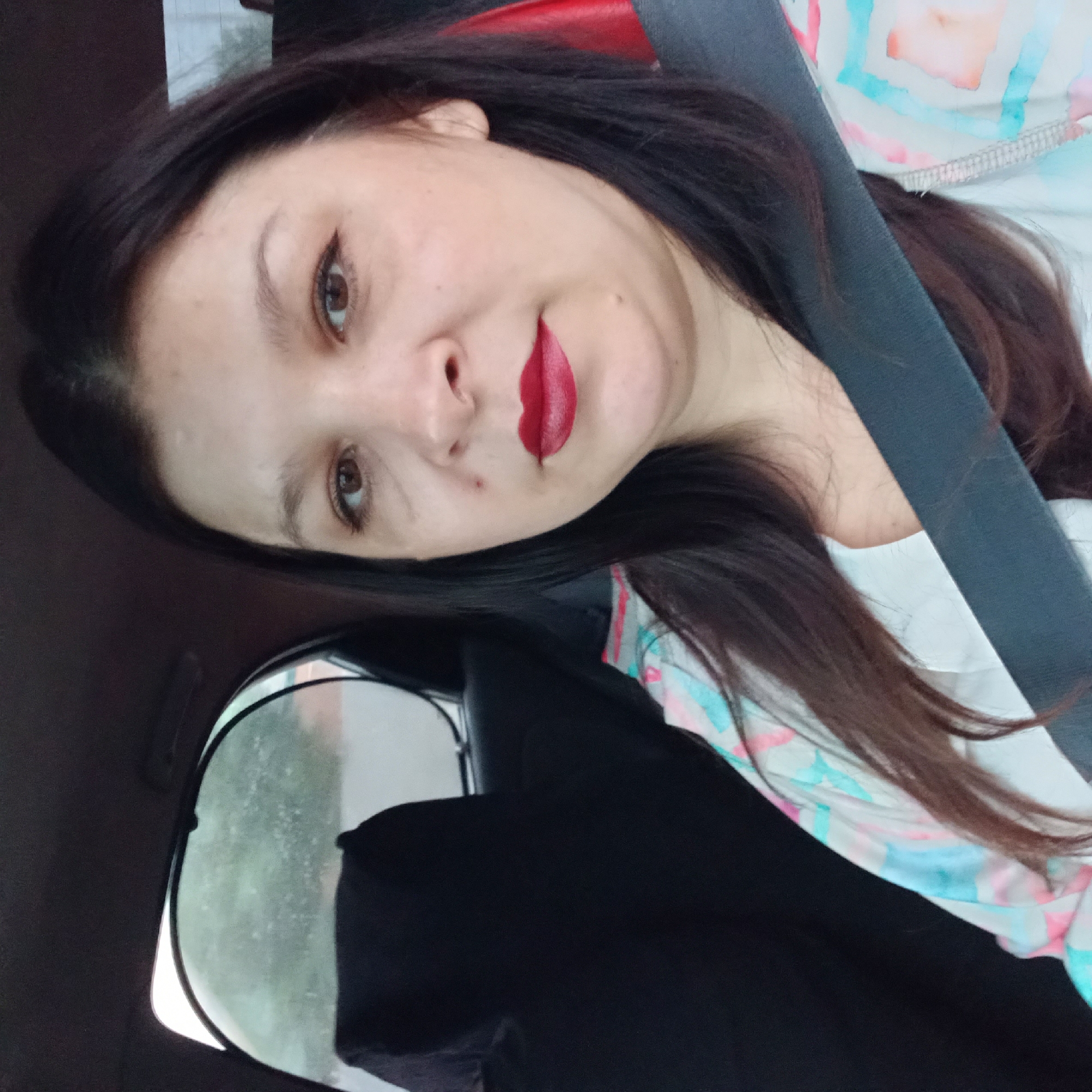 A woman with dark hair and red lipstick is sitting in the passenger seat of a car. She is wearing a white shirt with a colorful patterned design. A seatbelt is across her chest. The background shows a car seat and a sunshade on the window. The image is a selfie taken inside the car.