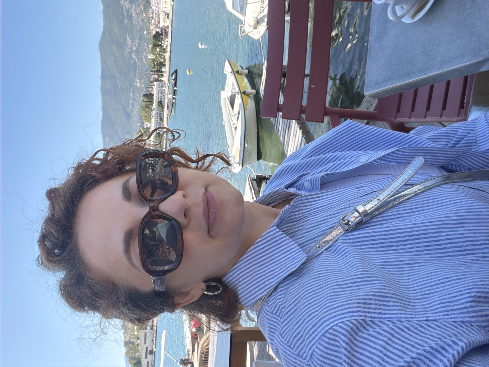 A woman with curly brown hair and dark tortoiseshell sunglasses is pictured in a close-up, outdoors, on a bright, sunny day. She is wearing a light blue and white striped button-up shirt with a silver strap, possibly from a purse or belt, visible across her chest. A silver hoop earring is visible on her left ear. Her expression is neutral, and her lips are slightly parted.
She is seated at a light gray table, with a white cup visible on its surface, and a red slatted chair beside her. The setting is a waterfront cafe or restaurant, overlooking a calm body of water. Several boats are moored in the blue-green water, including a prominent white boat with yellow trim, and another white boat with the Greek text "ΣΕΡΦΩ" (Serfo) painted on its bow. A wooden dock is also visible.
In the background, across the water, a picturesque coastal town with light-colored buildings stretches along the base of large, verdant mountains under a clear blue sky. The general location, indicated by the mountainous coastal landscape and the Greek text, is Poros, Greece, as specified in the context. The time of day appears to be daytime, likely morning or afternoon, given the bright sunlight. The overall atmosphere conveyed is relaxed and peaceful.
