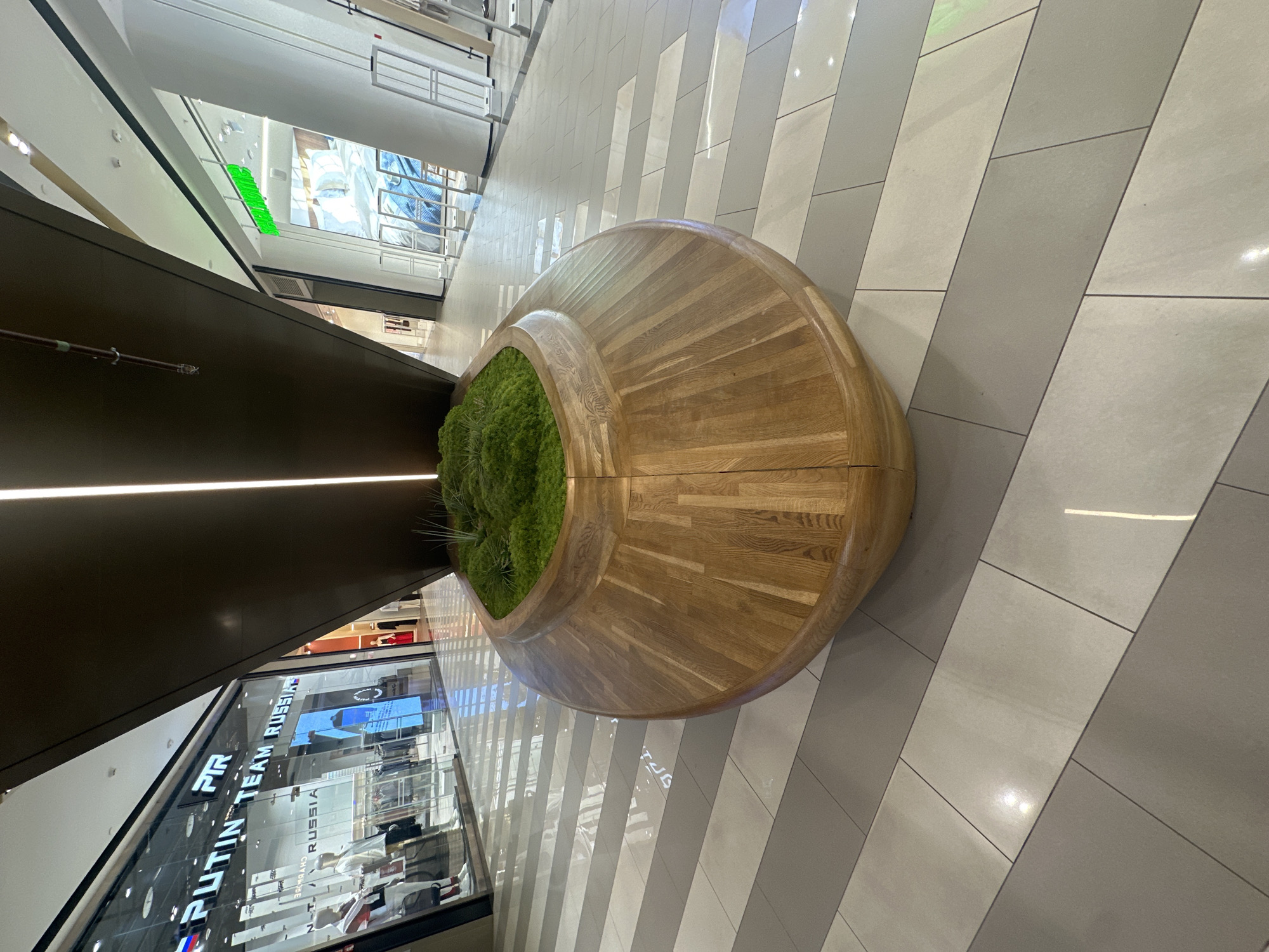 The image shows a high-angle view within a modern shopping mall or commercial space, likely in Родниковское сельское поселение, Ukraine. A large, circular wooden bench with a central patch of dense green plants serves as a focal point. The bench sits on a tiled floor with a diagonally striped pattern in shades of gray. Reflected light from the surrounding shops adds highlights to the tile surfaces.
In the background, there are storefronts featuring brightly lit interiors with visible displays of merchandise and branding, including the words "PUTIN TEAM RUSSIA." The lighting suggests a brightly illuminated indoor environment. The overall perspective gives a sense of the mall's layout and its integration of seating and green space within the shopping area. There are no people directly visible in the immediate foreground, but reflections in the glass storefronts suggest their presence.