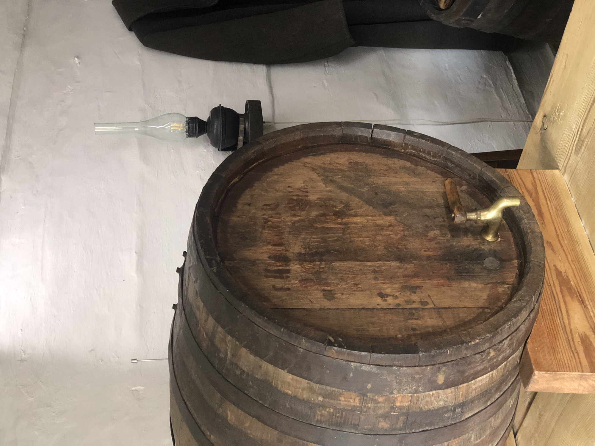 A wooden barrel with metal hoops sits on a wooden support. A brass spigot is attached to the front of the barrel. To the right of the barrel, a black oil lamp with a glass chimney sits on a wall-mounted shelf. A dark garment, possibly a coat, hangs on the wall to the right of the lamp. The background is a white, textured wall. The scene appears to be indoors, likely in a historical setting in Shushenskoye, Russia, given the context of the location.