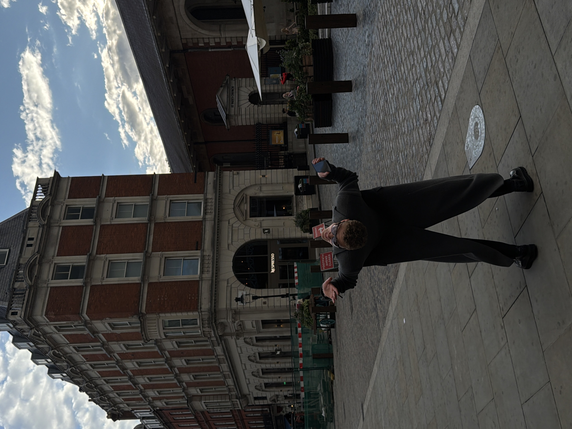 A full shot shows a person bending backward on a cobblestone street in front of a large brick building in London, United Kingdom. The person is wearing a dark gray suit and is holding a phone in their right hand. The building behind them is multi-story with numerous windows and architectural details. In the background, there is another building with a sign that reads "ST. PAUL'S CHURCH." The sky is partly cloudy with blue sky visible. The street is lined with planters and bollards. There is a "FOOTPATH AHEAD CLOSED" sign visible. The scene appears to be a public square or street.