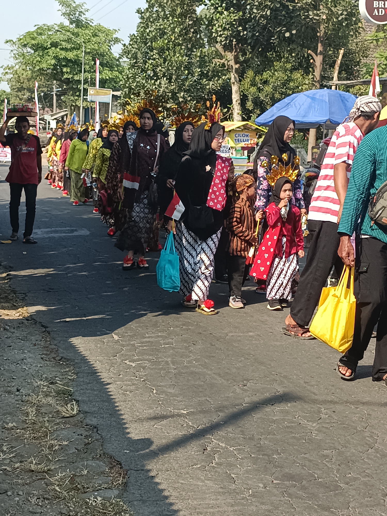A group of people, including adults and children, are participating in a procession on a paved road in Jatitengah, Indonesia. Many individuals are wearing traditional Indonesian attire, characterized by batik fabrics, headscarves (hijabs), and head coverings. Some participants are carrying small Indonesian flags. The environment appears to be a sunny day with trees lining the roadside and buildings visible in the background. A blue umbrella is set up on the right side of the frame. The road surface is cracked and worn, with some sparse vegetation growing along the edge. The people are walking in a line, with some carrying items like bags.