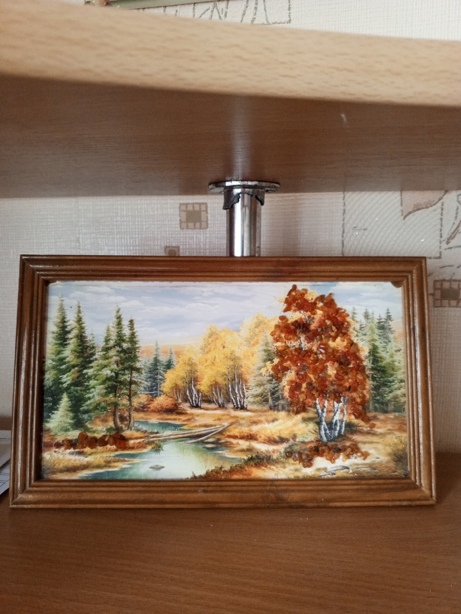 A framed painting of an autumn forest scene is displayed on a shelf. The painting depicts a tranquil landscape with a winding river flowing through a forest of evergreen and deciduous trees. The deciduous trees are ablaze with the warm colors of autumn, featuring vibrant yellows and oranges. One prominent tree in the foreground has fiery orange leaves. The river reflects the sky and surrounding foliage. A rustic wooden bridge, made of fallen logs, spans the water. The overall scene evokes a sense of peace and natural beauty. The painting is housed in a simple wooden frame and is positioned on a wooden shelf. The background behind the painting appears to be a textured wallpaper. There is no visible text in the image. The lighting suggests it is daytime, with soft, natural light illuminating the scene.