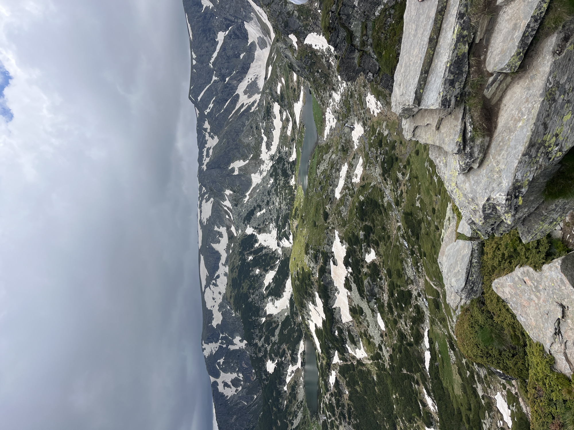 The image depicts a panoramic view of a mountainous landscape with patches of snow. In the foreground, large, grey rocks are visible, some covered in green lichen. The midground features a valley with a dark, reflective lake and rolling green hills interspersed with snow. Further in the background, a rugged mountain range with snow-capped peaks dominates the scene. The sky is overcast with thick, grey clouds. The location is identified as the Rila Mountains near Panichishte, Bulgaria, a popular hiking area known for its glacial lakes, such as the Seven Rila Lakes, which this scene likely represents.