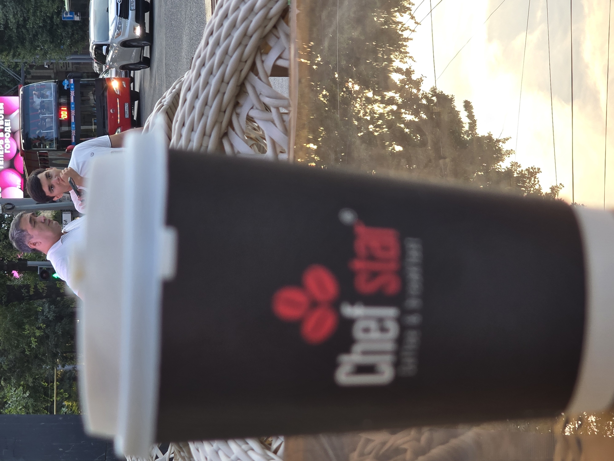 An outdoor scene in Shymkent, Kazakhstan, is captured with a close-up of a black coffee cup in the foreground. The cup has a white lid and features a red logo with three coffee beans and the text "Chef star coffee & breakfast". Behind the cup, a wicker chair is visible. Further in the background, two men are standing on a city street, with one in a white polo shirt and the other in a white t-shirt. A red double-decker bus and a white SUV are on the street. The sky is visible with reflections of trees and sunlight on a wet surface, possibly a table.