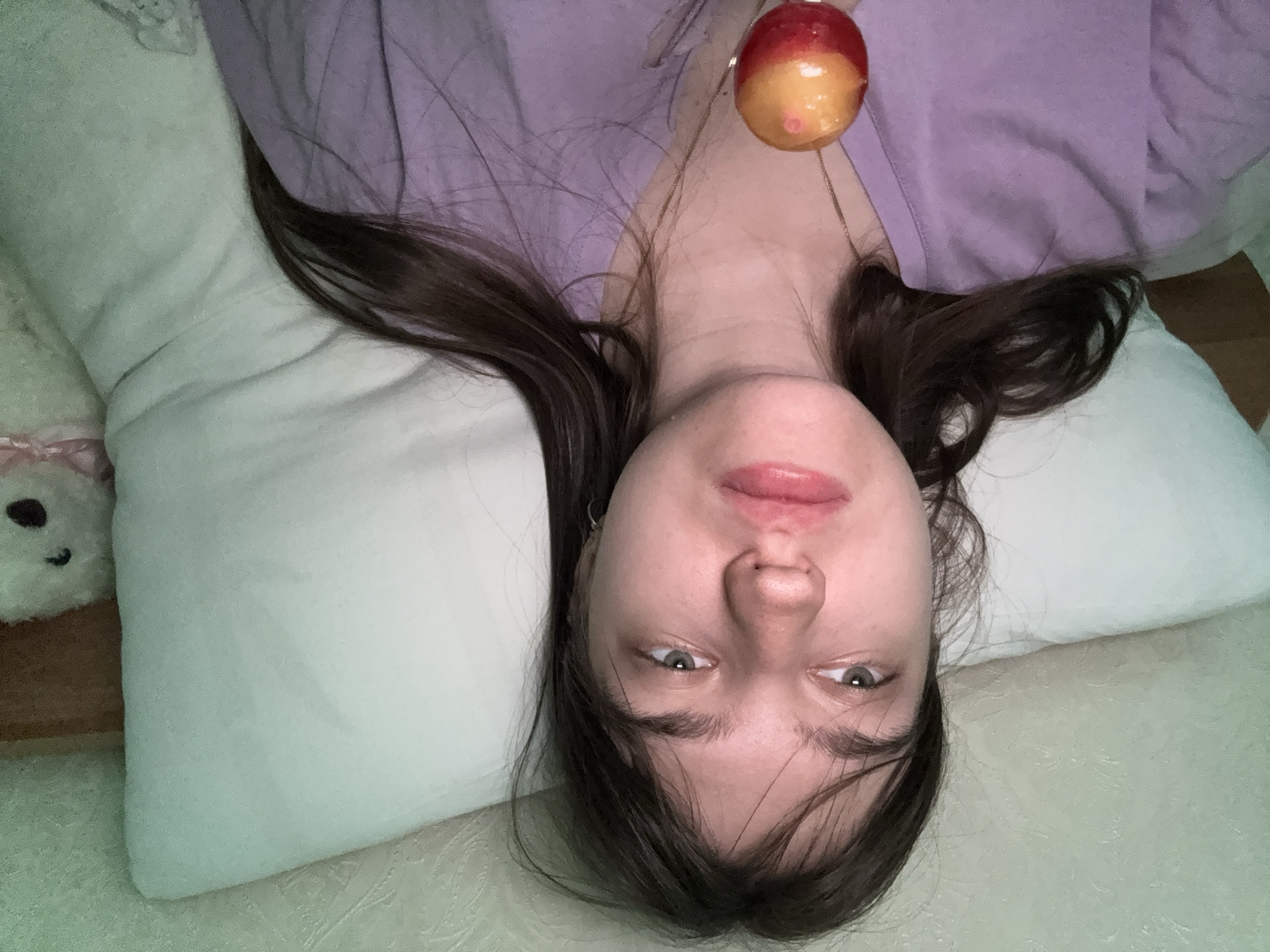 A female person with brown hair is lying down on a white pillow, wearing a lilac-colored garment and a necklace with an apple-shaped pendant. A white stuffed animal is partially visible in the upper left corner of the image. The location is Unknown, Unknown.