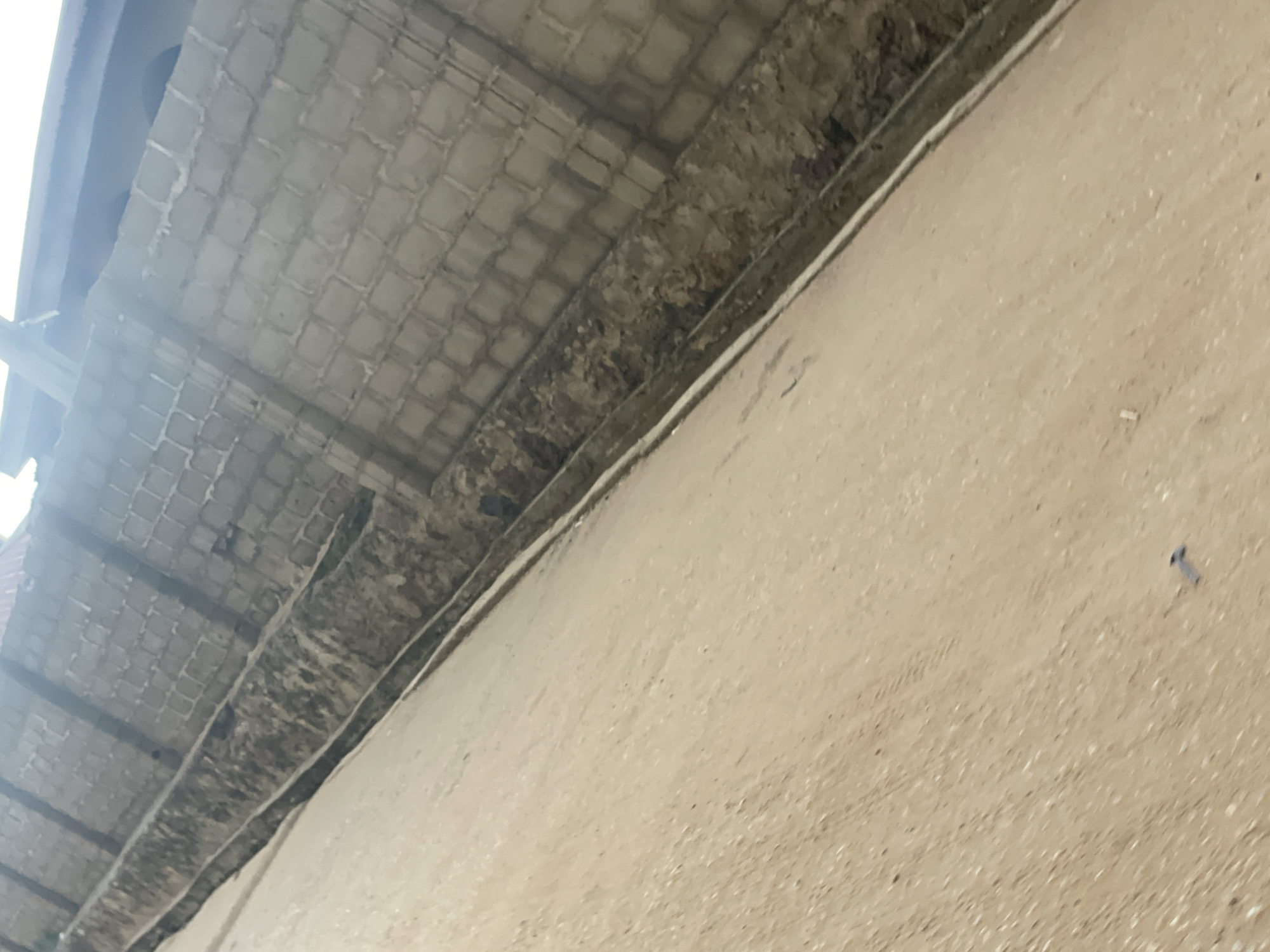 The image shows a side view of a drainage channel along a brick wall, in what appears to be an urban environment in Lokoja, Nigeria. The drainage channel is partly filled with dirt and debris. The brick wall is constructed from light-colored rectangular blocks and has a rough texture. Above the wall, a section of a roof is visible, and some blurry figures can be seen. The ground in front of the drainage is made of sand or loose soil, and there are some small objects scattered on it, including what looks like a screw. The lighting suggests it is daytime.