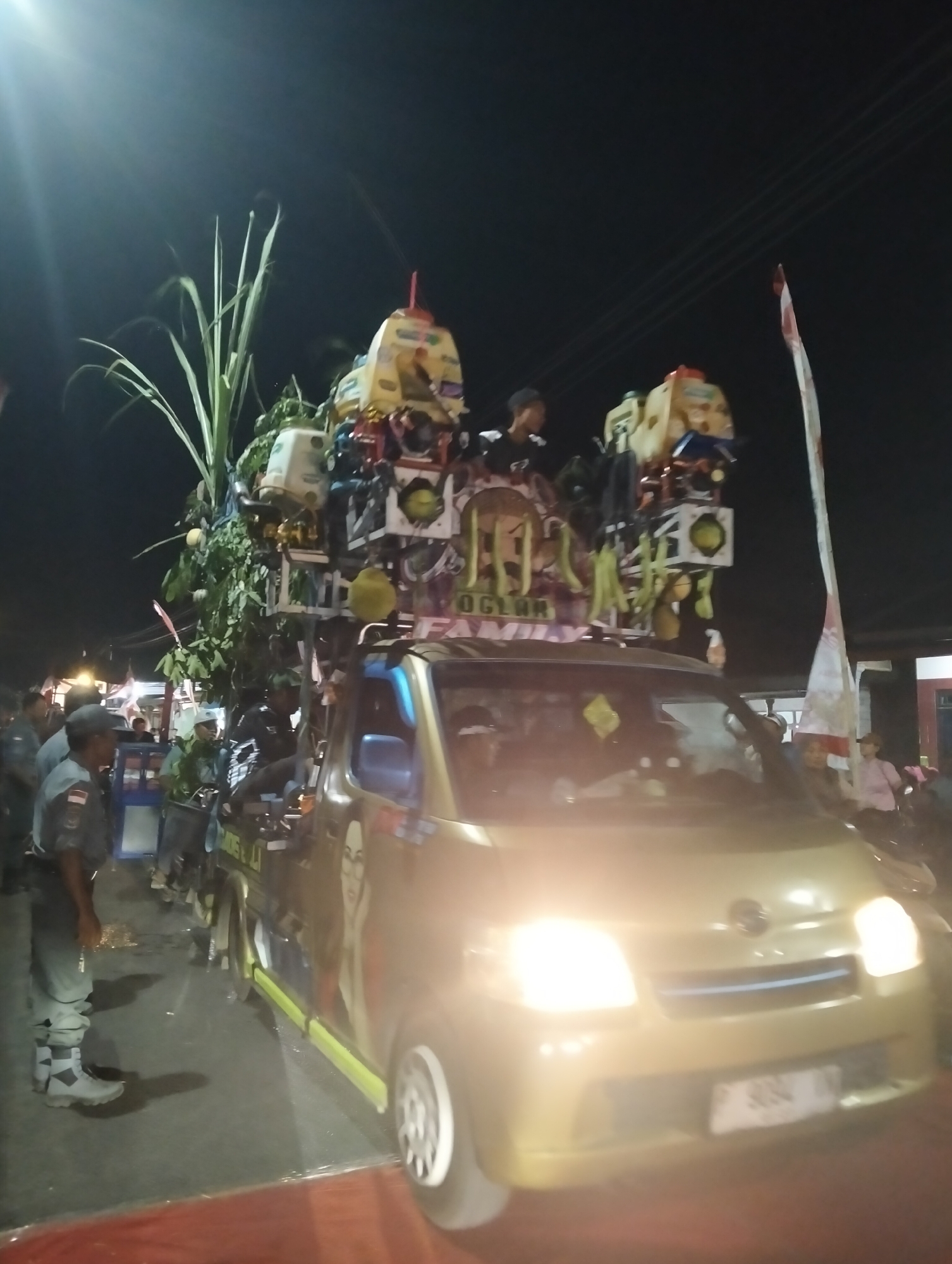 A golden minivan decorated with a large, colorful, multi-tiered structure, likely for a parade or celebration, is parked on a paved road at night. The structure on the van's roof is adorned with various objects, including what appear to be speakers, lights, and greenery, possibly sugar cane stalks and fruit. A woman's portrait is visible on the side of the minivan. Several people are standing around the vehicle. On the left, three individuals in dark uniforms with grey pants and caps are standing on the road. One of them is looking towards the van. In the background, a red and white Indonesian flag is mounted on a pole. The environment suggests a street scene in Sumbersewu, Indonesia, given the presence of the Indonesian flag and the style of the vehicle and decorations. The overall lighting indicates it is nighttime, with the minivan's headlights and some artificial lighting illuminating the scene.