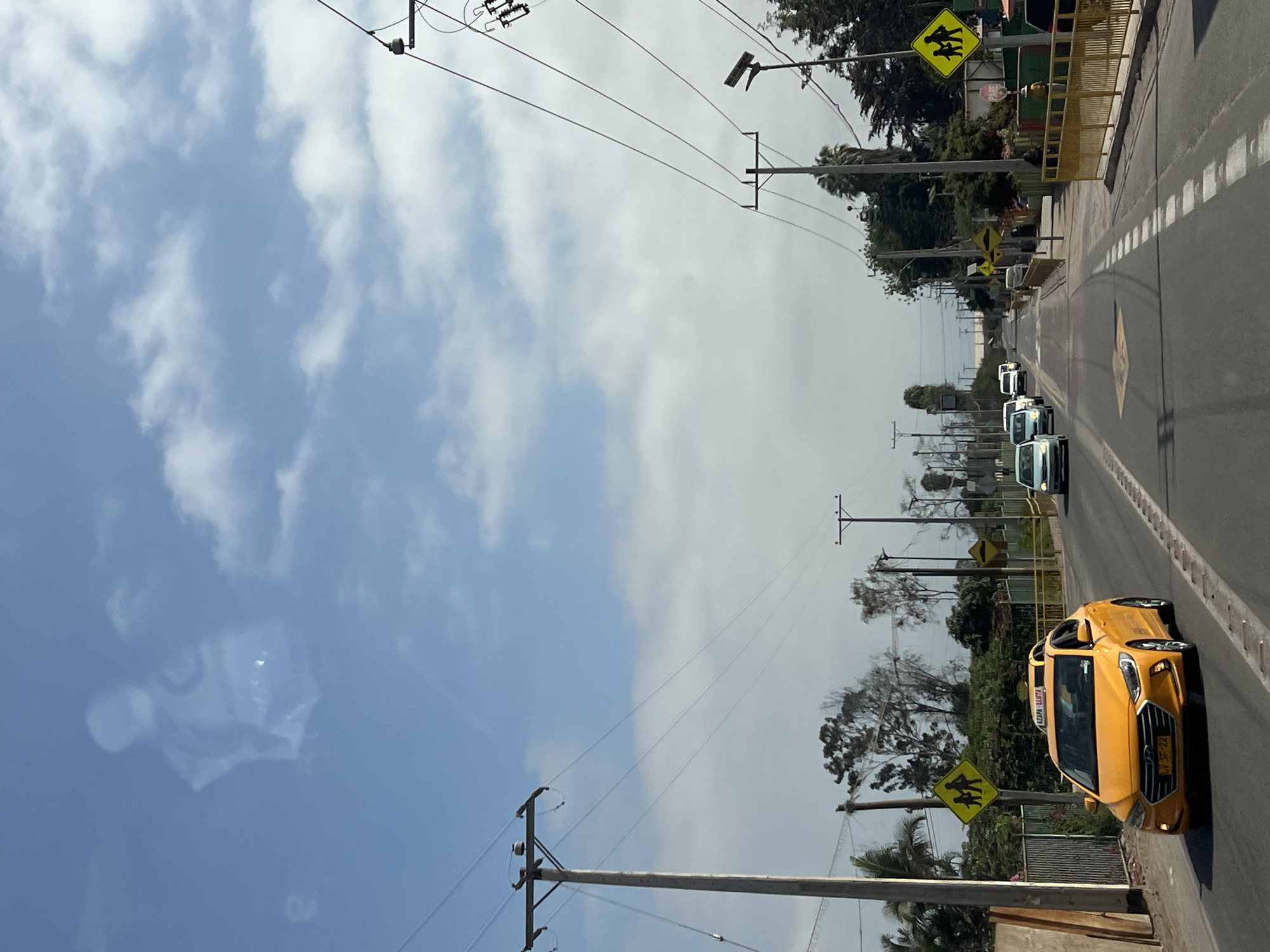 The image depicts a street scene in Arica, Chile. A yellow taxi is prominent in the foreground, driving on the right side of the road. Further down the road are several other cars and a white van. Utility poles and wires run along both sides of the street. School crossing signs are visible. The sky is partly cloudy, with blue patches. The overall environment appears to be a typical urban or suburban road in Arica.