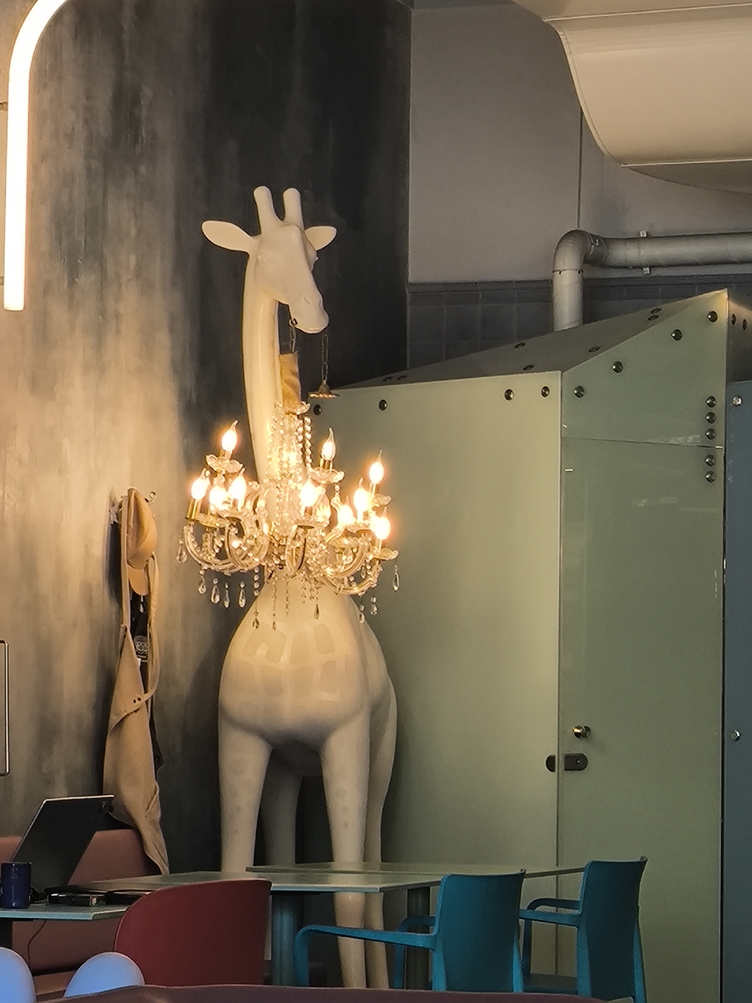 The image depicts an interior space with quirky decor, featuring a large white giraffe statue adorned with a lit chandelier around its neck. The giraffe stands next to a table with blue chairs, suggesting a dining area. To the left, a wall with a light fixture and an apron hanging adds to the scene's eccentric character. The area seems to be enclosed by partition walls, one of which has a door. The location is in the city of Unknown, Unknown.