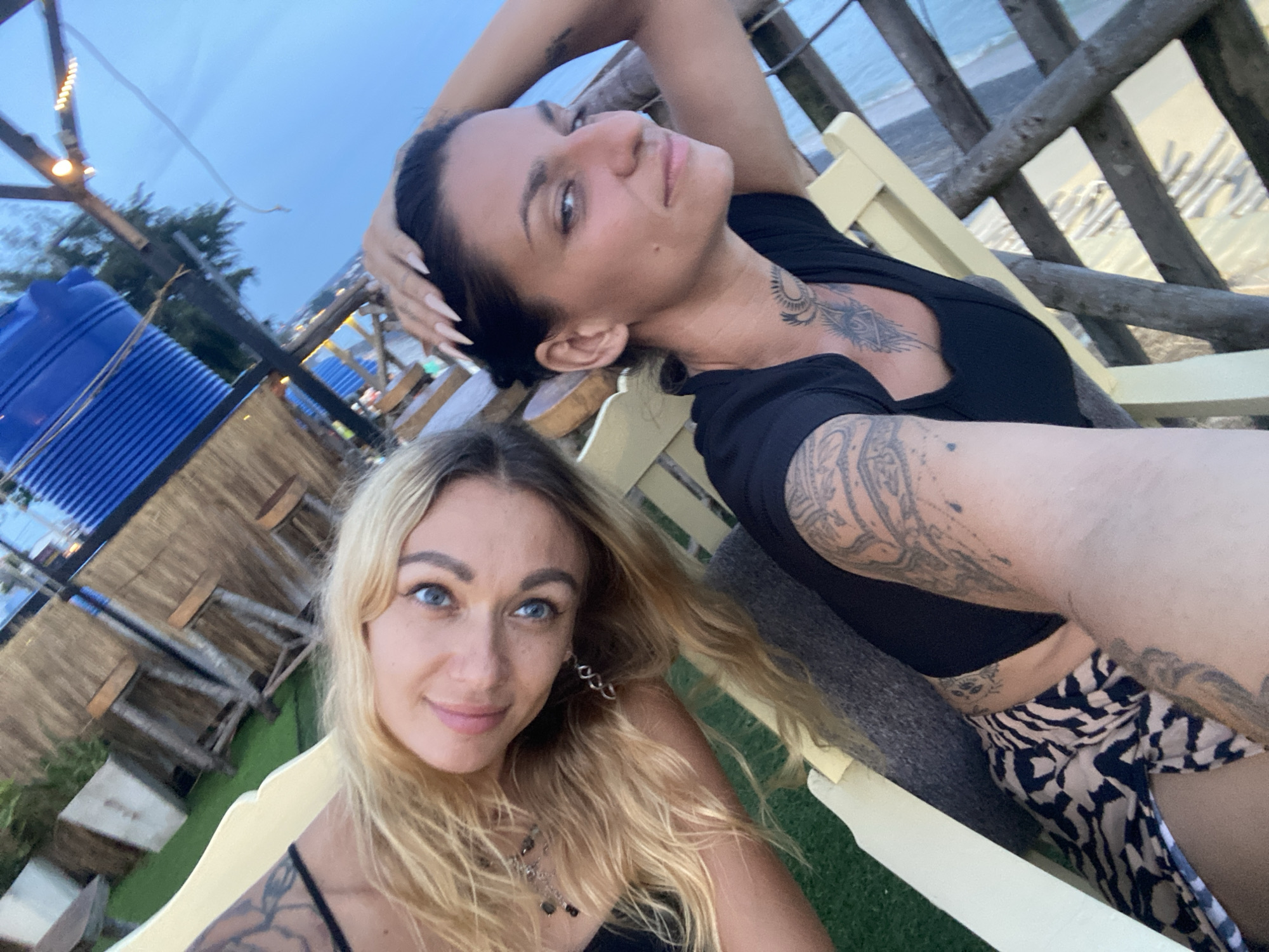 Two women are in a selfie taken outdoors in Phu Thuy Ward, Vietnam. The woman on the left has long blonde hair, blue eyes, and is wearing a black top and a leopard print skirt. She has tattoos on her left arm and chest. The woman on the right has dark hair, brown eyes, and is wearing a black top. She has tattoos on her arm and chest. They are sitting on chairs in front of a bamboo fence and a blue water tank. The sky is blue and it appears to be late afternoon or early evening.