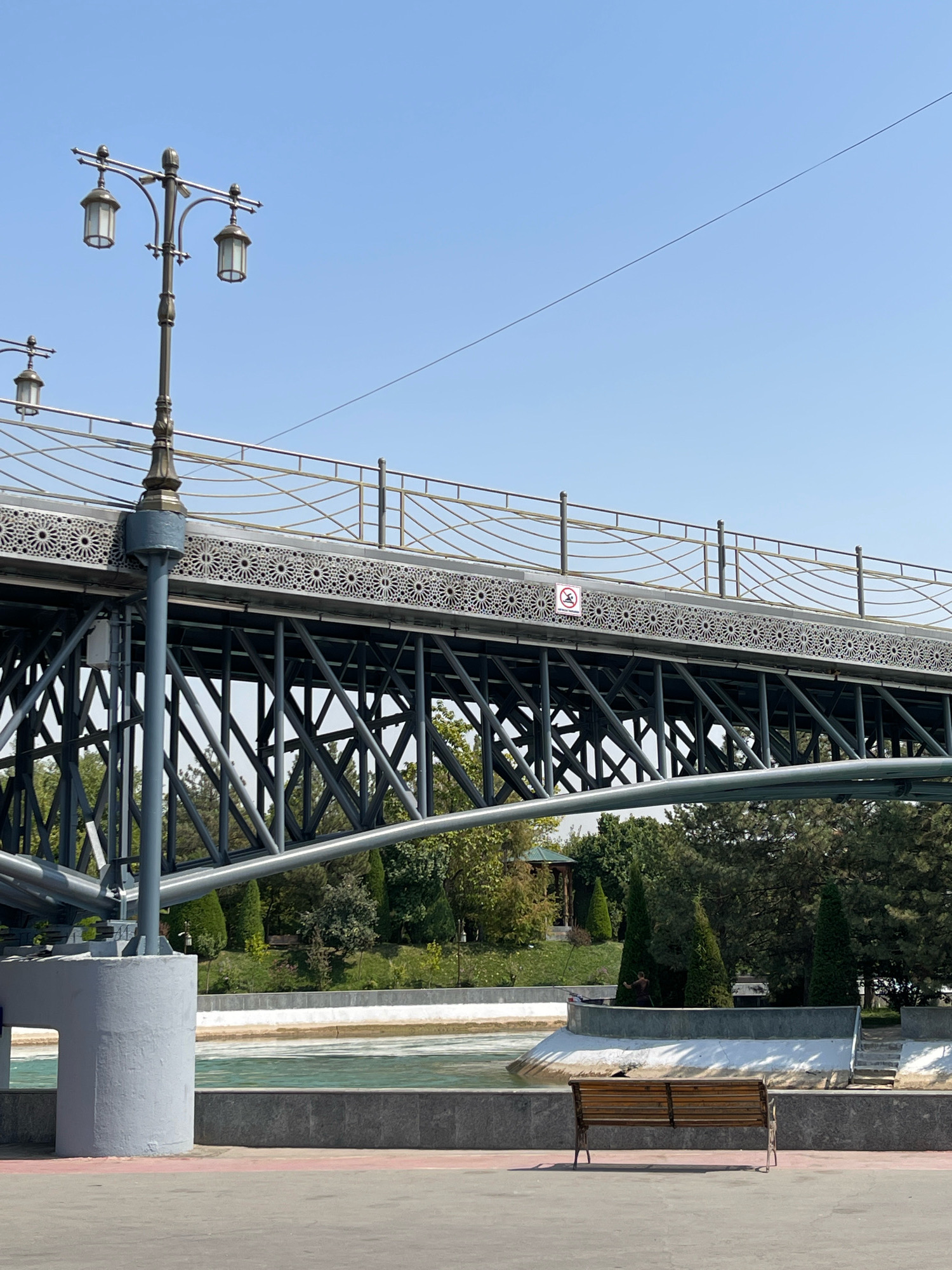 The image depicts the "Bridge of Sighs" (Ko'z yosh ko'prigi) located in Tashkent, Uzbekistan, situated above the Ankhor Canal. The bridge is a prominent architectural feature, characterized by its dark metal structure with intricate, ornamental patterns along its sides. A double-headed street lamp with a vintage design stands to the left of the bridge, casting light on the scene. Below the bridge, the Ankhor Canal flows with turquoise-colored water, bordered by a concrete walkway. On the walkway, a simple wooden bench is positioned in the foreground, facing the canal. Behind the bridge, lush green trees and manicured shrubs create a park-like setting. The sky above is a clear, bright blue. No individuals are visible in the image.