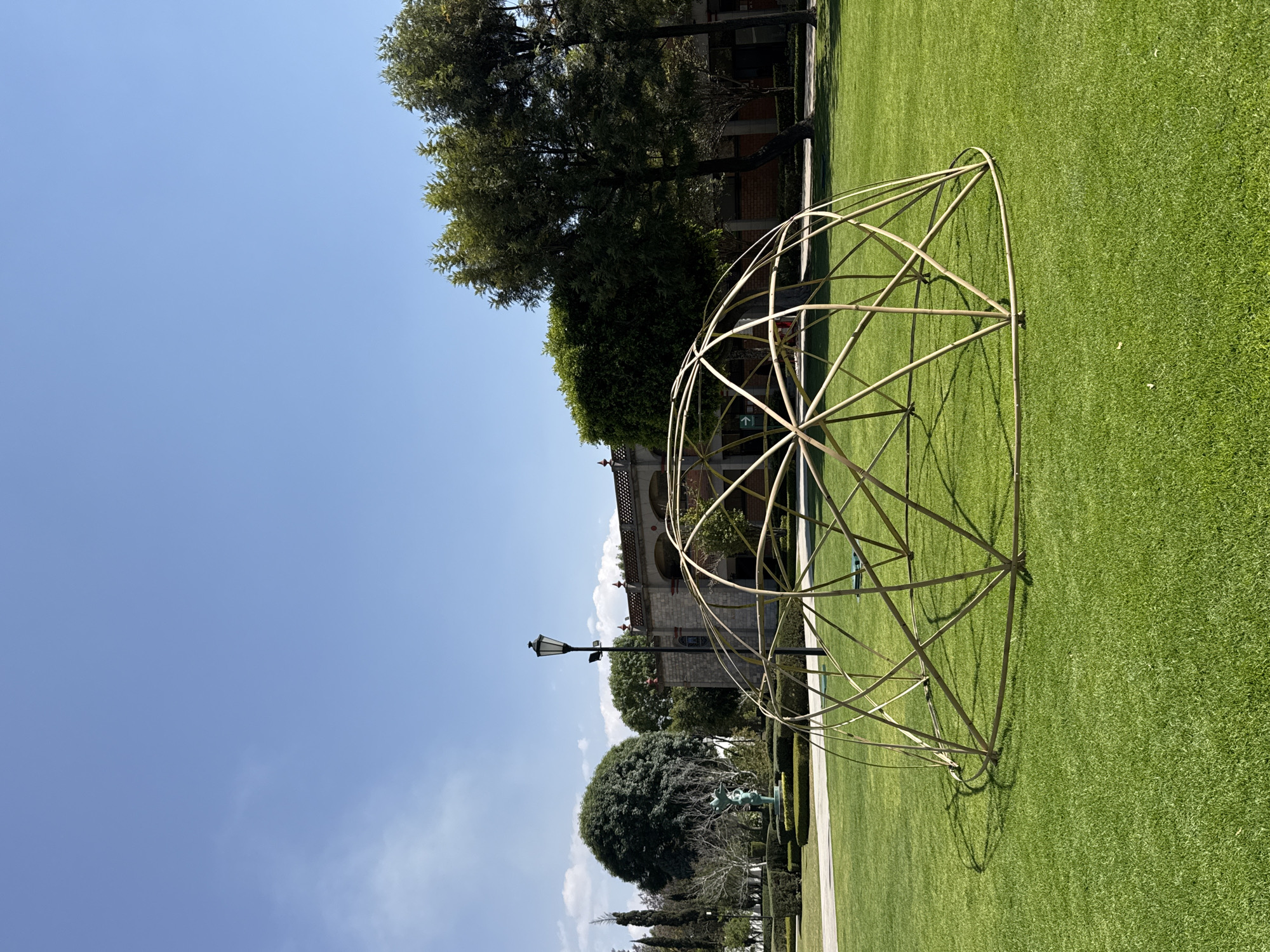 The image displays an outdoor scene in San Andrés Cholula, Mexico, under a bright blue sky with a few scattered white clouds. A large, partially assembled geodesic dome structure, constructed from light-colored tubular elements, occupies the foreground, resting on a manicured green lawn and casting a distinct shadow to its left. In the midground, a black, ornate streetlamp with two lights stands upright. The background features a traditional-style building with a reddish-brown base, light-colored upper sections, arched openings, and a tiled roof adorned with decorative elements. To the left of the building, a green statue depicting two figures is situated amidst neatly trimmed hedges and a paved pathway. The environment includes various trees, such as large rounded deciduous trees and taller evergreen varieties, along with dense shrubbery. No people are visible within the scene, and no specific actions or interactions are occurring.