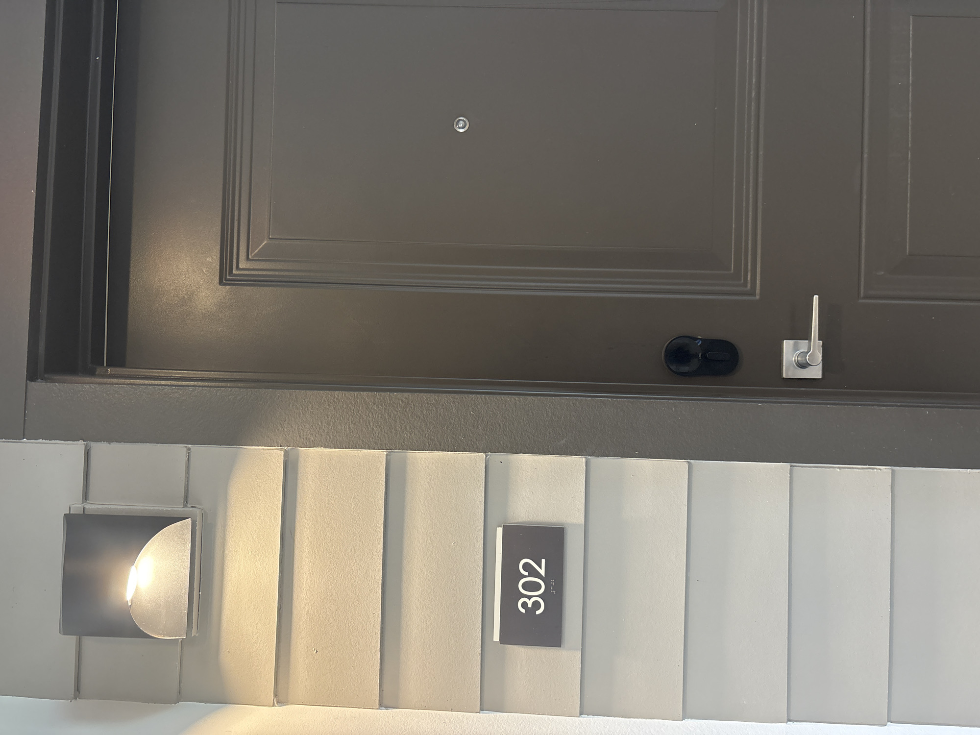 A dark gray, paneled apartment door is visible on the right side of the image, featuring a silver doorknob, a smart lock, and a peephole. To the left of the door, on a light-colored, horizontally-paneled wall, is a dark rectangular sign displaying the number "302" in white text, with accompanying braille. Above the sign, a modern, square outdoor light fixture with a curved diffuser illuminates the wall. The scene is likely in an apartment building hallway or exterior.