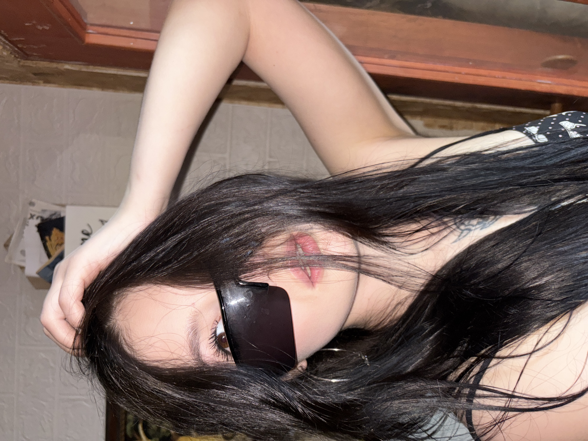 A woman with long, dark hair is lying on her back with her left hand near her head and her right hand behind her. She is wearing dark sunglasses that obscure one eye, and a black top with a white polka dot pattern. The background includes a tiled wall, what appears to be a cabinet, and a white object with writing on it. The image doesn't contain enough information to determine the exact location or city.